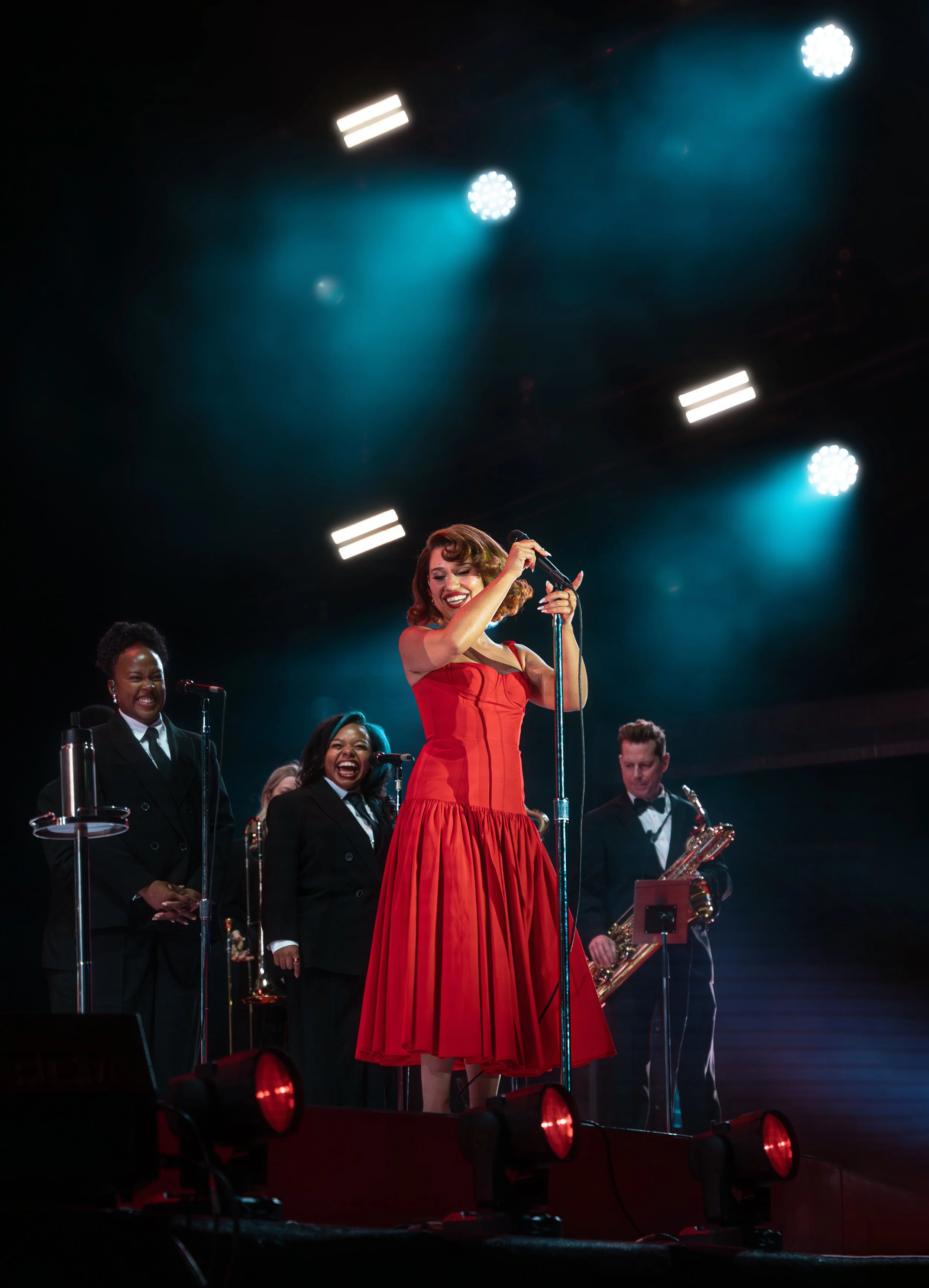 A woman in a red dress singing into a microphone on stage, surrounded by backup singers and a saxophonist, under stage lights.