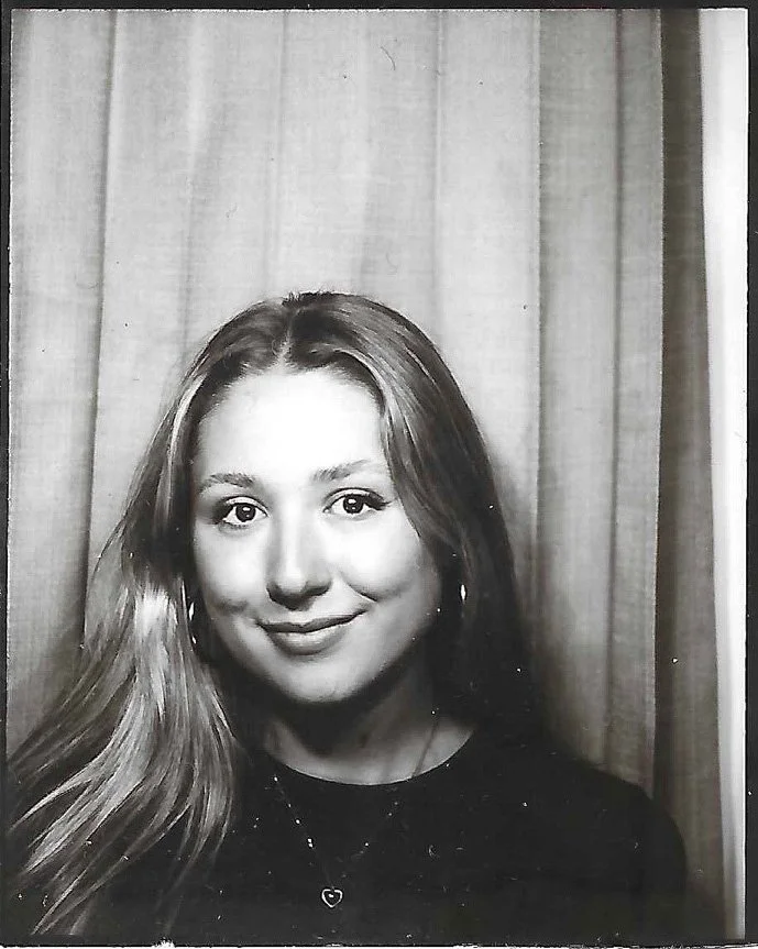 Portrait of a young woman with long hair, smiling, wearing a black top and a necklace with a heart pendant, standing in front of curtains.