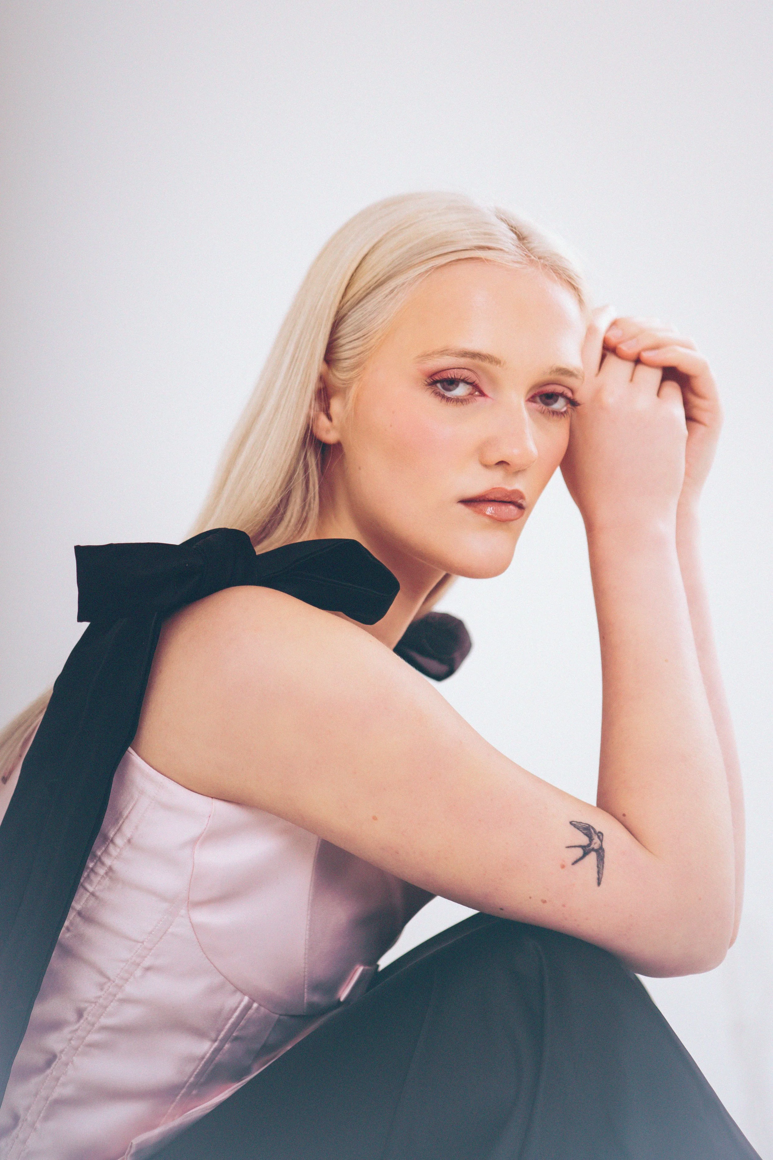 A woman with long blonde hair and light skin, wearing a pale pink top with black ribbon details, sits against a plain light background. She has a tattoo of a bird on her forearm and is resting her head on her hand, looking at the camera with a neutra