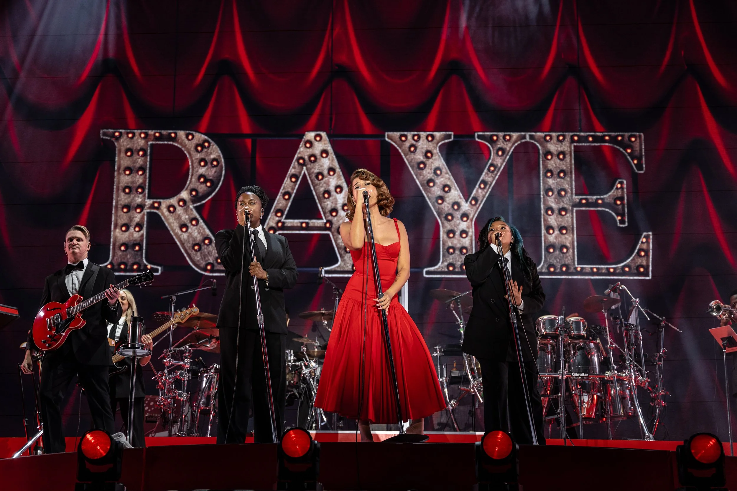 A musical band performs on stage with a large illuminated sign behind them that reads 'RAYE' in marquee lights. The band includes four members, two women and two men, singing and playing guitar and drums, with a red curtain backdrop.