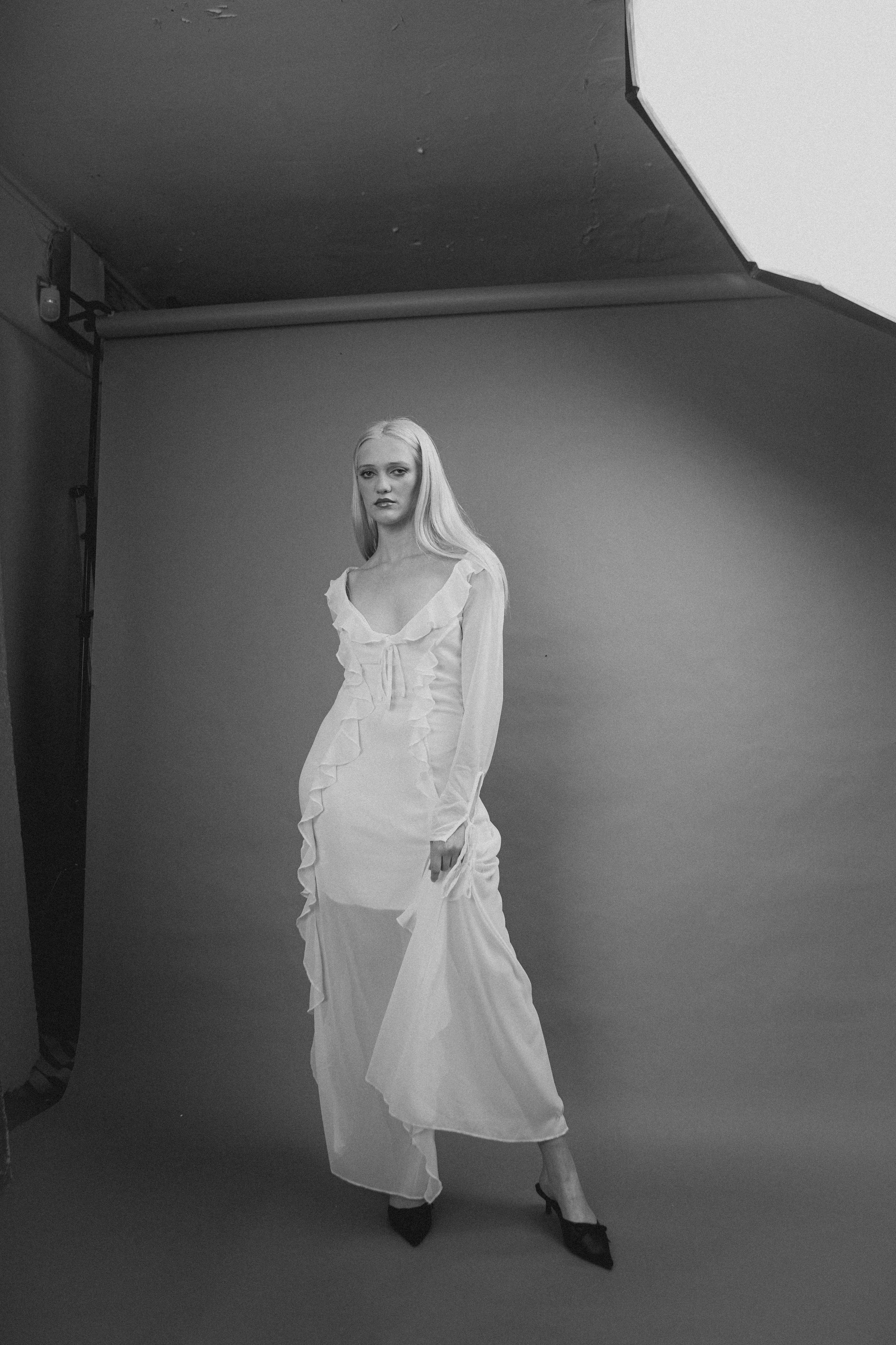 Black and white photo of a woman in a long, ruffled, vintage-style dress standing in a photography studio with a plain backdrop and studio lighting.