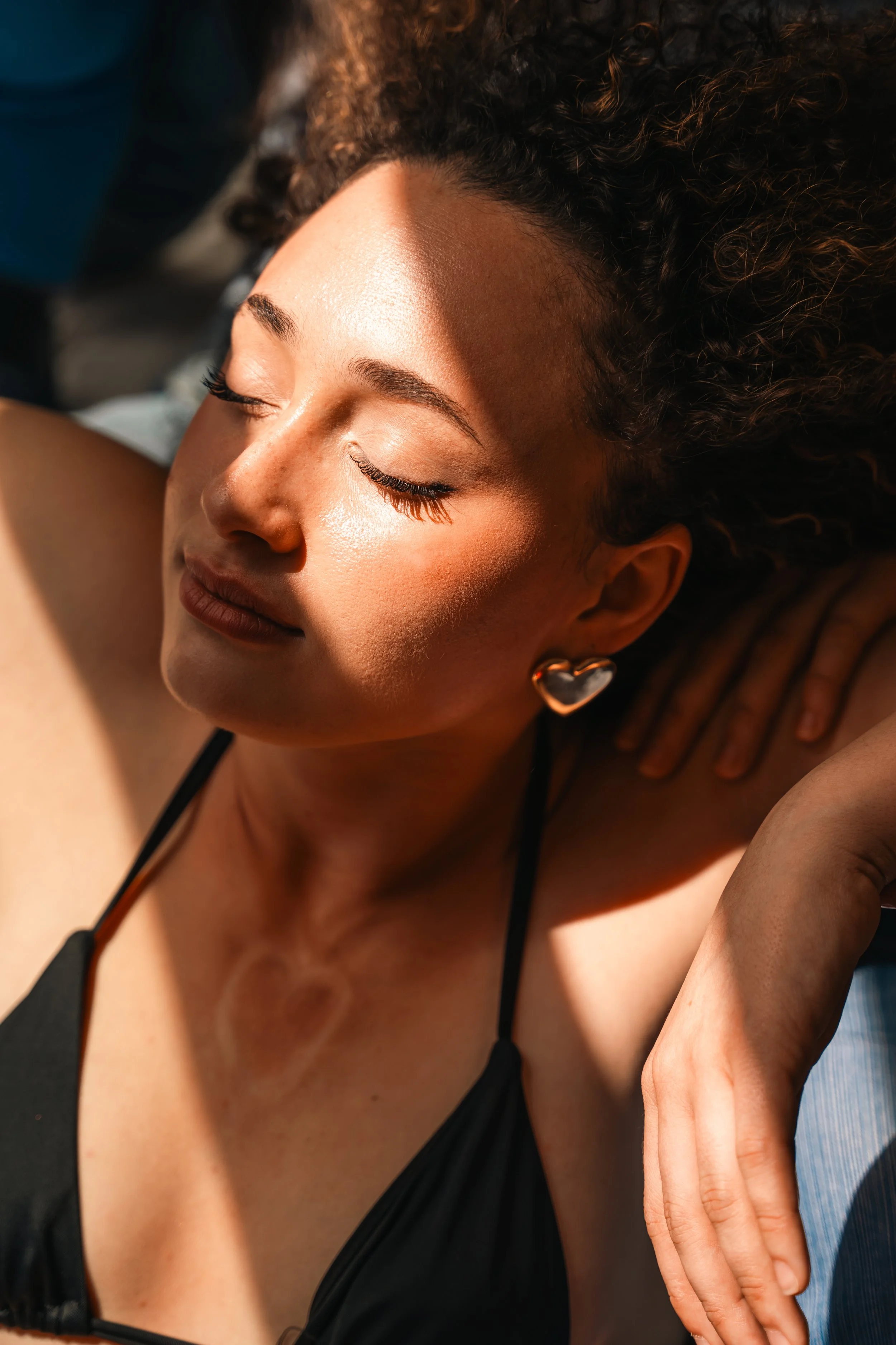 Close-up of a woman with curly hair, wearing heart-shaped earrings, with sunlight on her face and a black spaghetti strap top, resting with eyes closed.