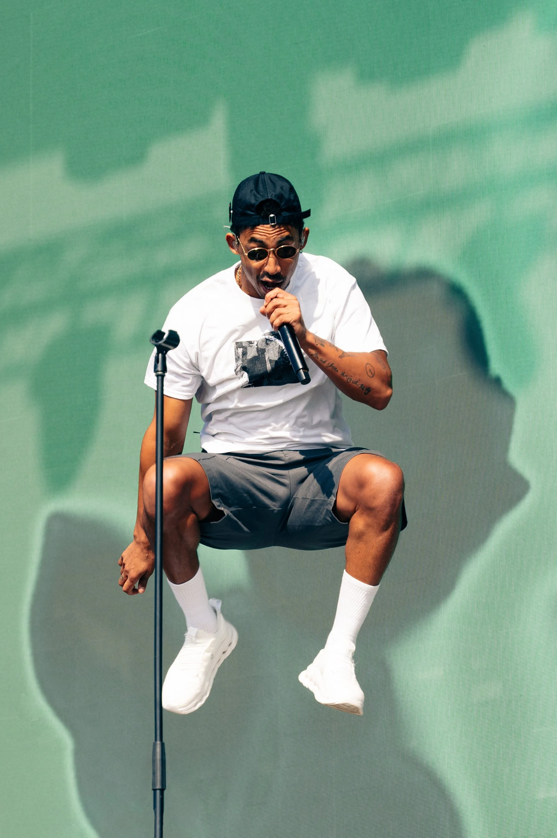 A man wearing sunglasses, a black cap, a white T-shirt, gray shorts, and white sneakers is sitting on a microphone stand, holding a microphone and singing or speaking with a large green abstract background.