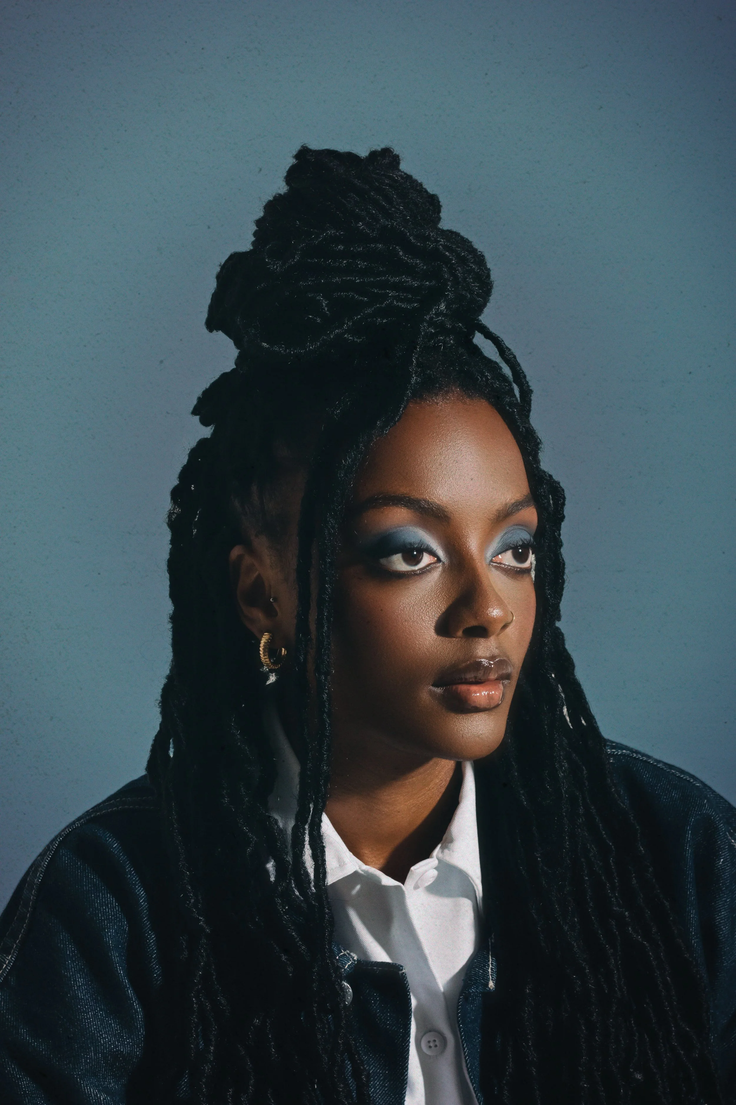 A woman with dark skin and long black dreadlocks styled in an updo, wearing gold hoop earrings, smoky eye makeup, and a leather jacket over a white shirt.