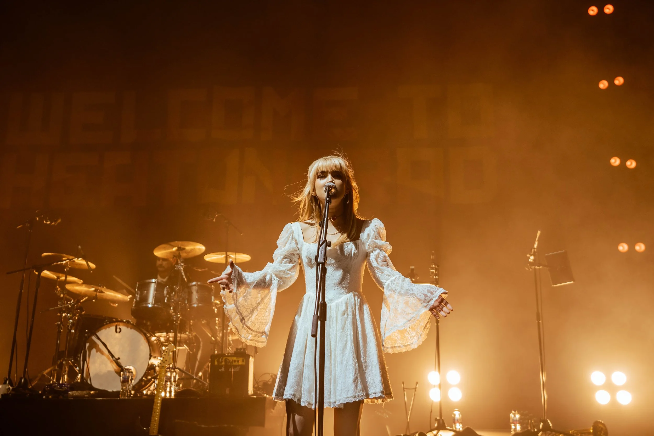 A woman singing on stage with a band, illuminated by warm stage lights.