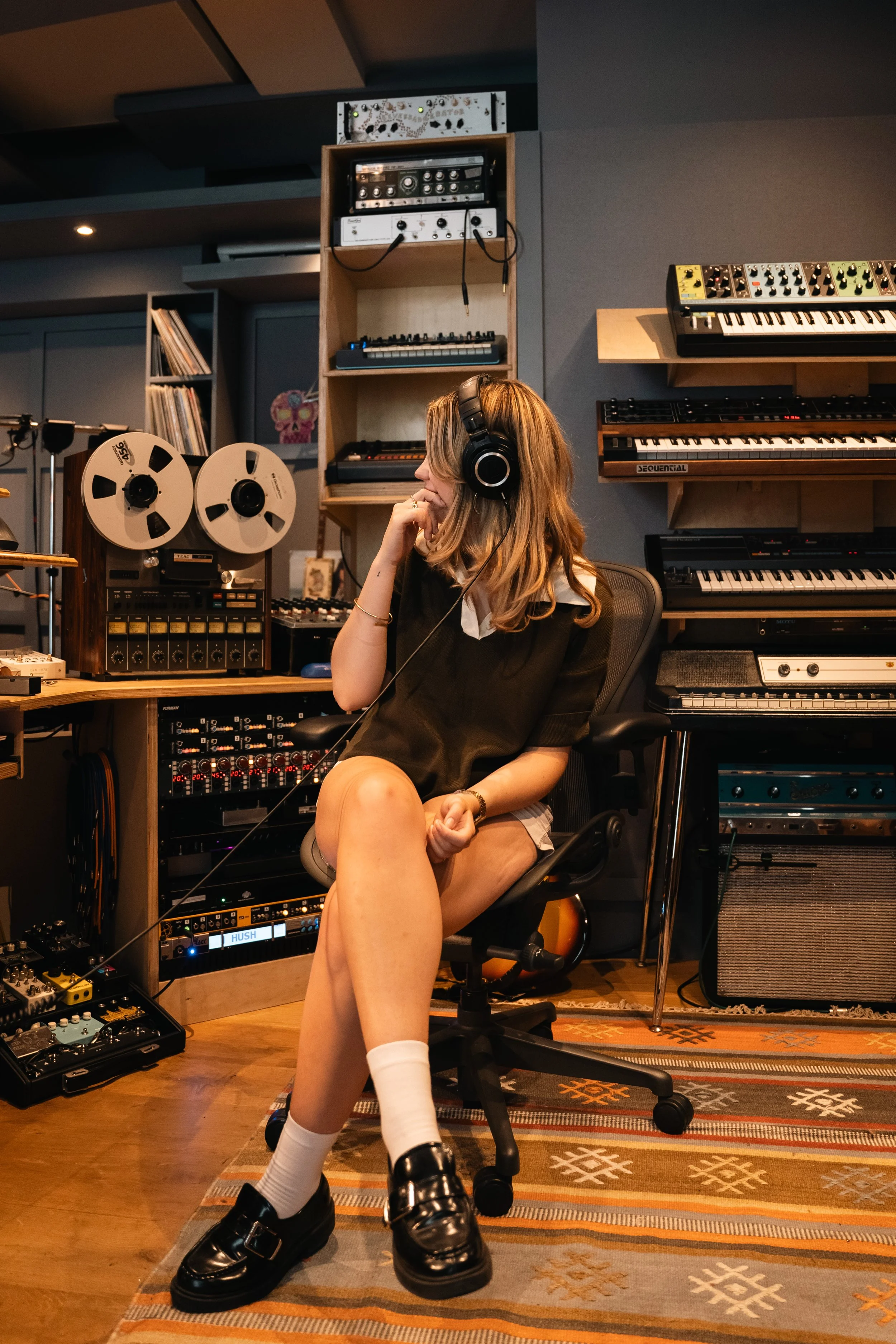 A young woman with headphones sitting in a recording studio, surrounded by music equipment and instruments.