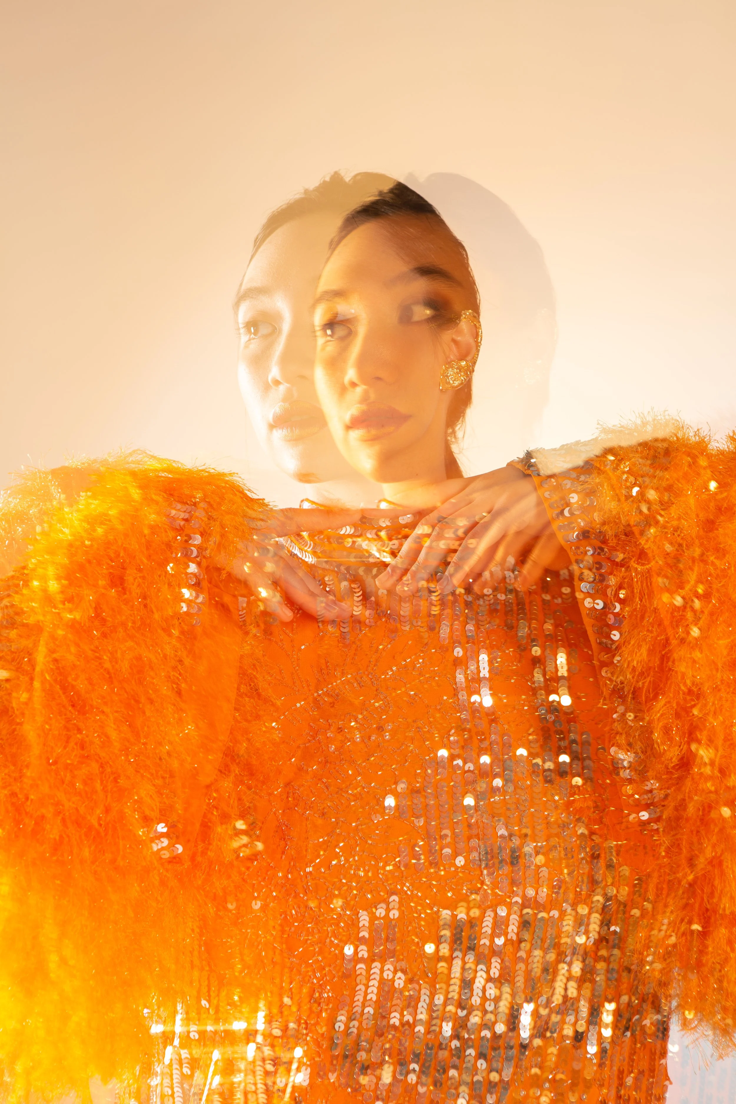 A woman wearing an orange sequined dress and matching fuzzy sleeves poses with hands near her neck against a gradient background, creating a double exposure effect.