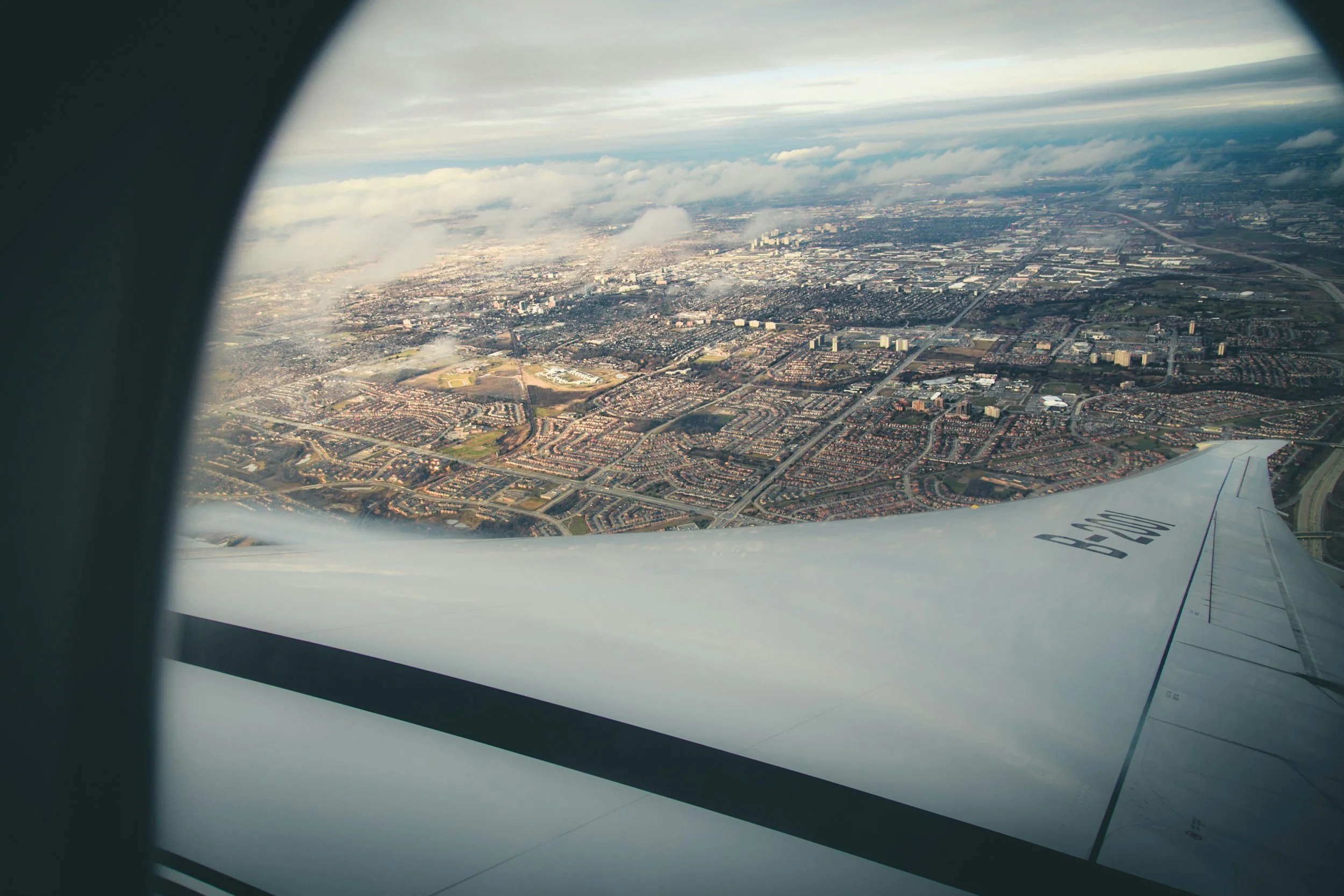View of a city from an airplane window, showing buildings, roads, and neighborhoods below, with part of the airplane wing visible in the foreground.