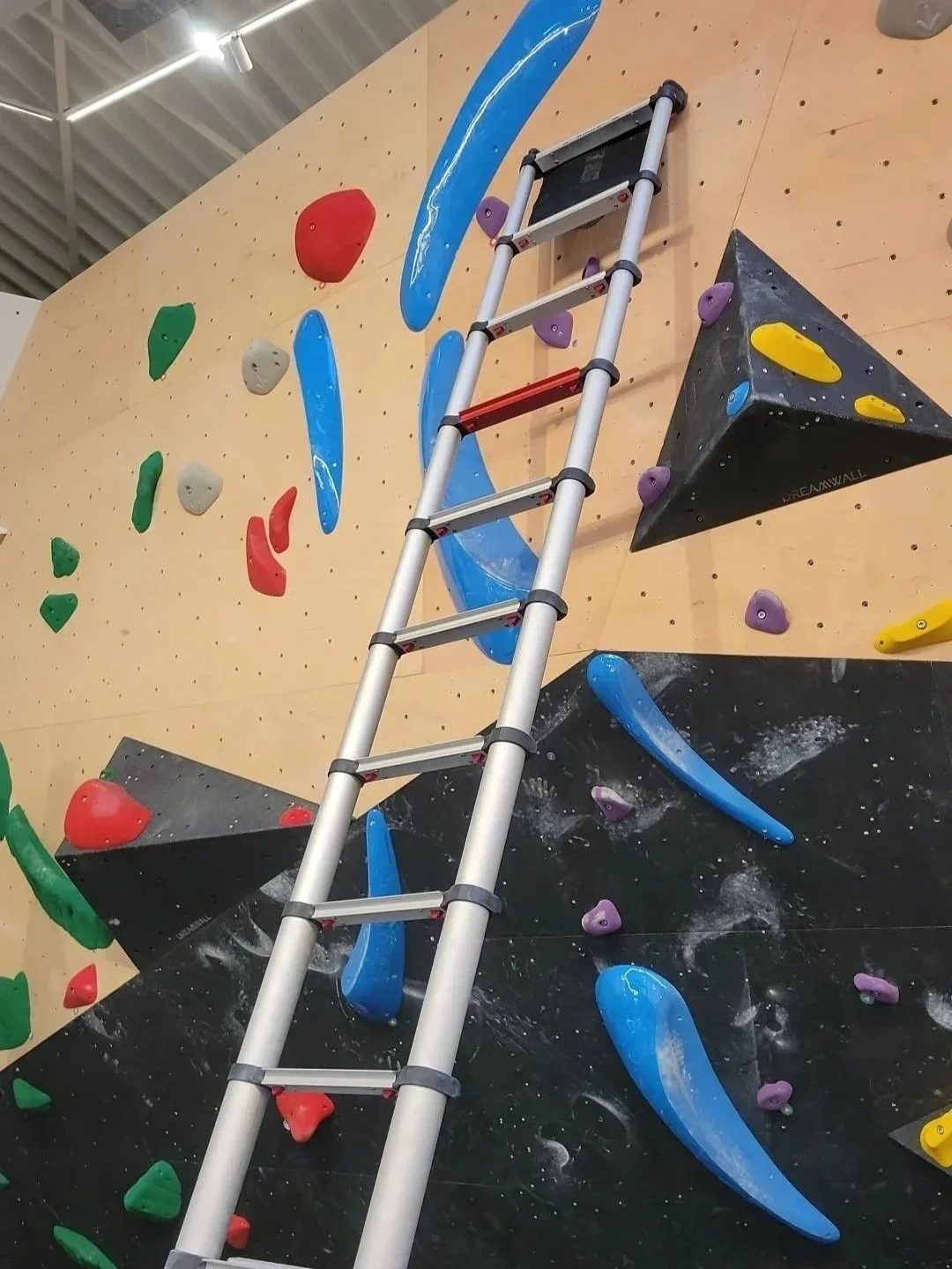 Indoor rock climbing wall with colorful holds and a ladder leaning against it.