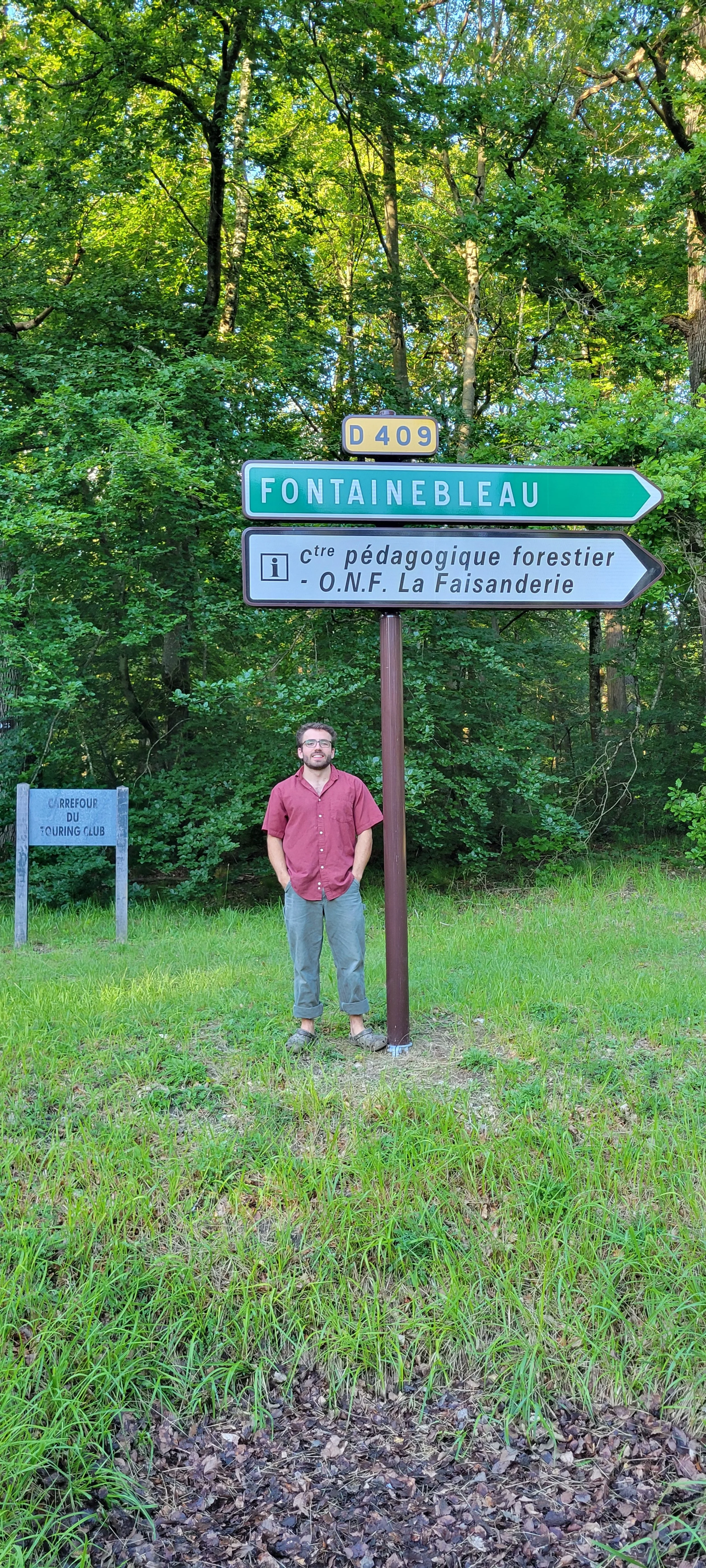 A man with glasses and a beard, wearing a red short-sleeved shirt and gray pants, standing outdoors on grass in front of a signpost. The signpost has signs pointing to Fontainebleau and a forestry education center with forest information, surrounded by lush green trees.