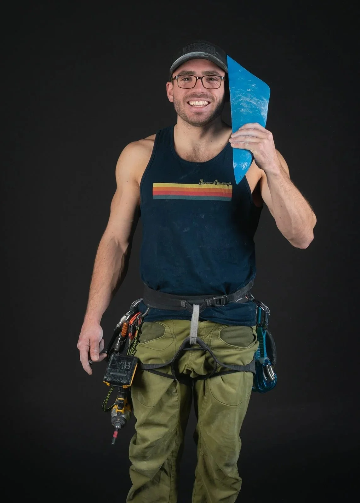 A man with glasses and a cap holding a blue climbing hold, dressed in climbing workout attire, with climbing gear around his waist, smiling against a black background.