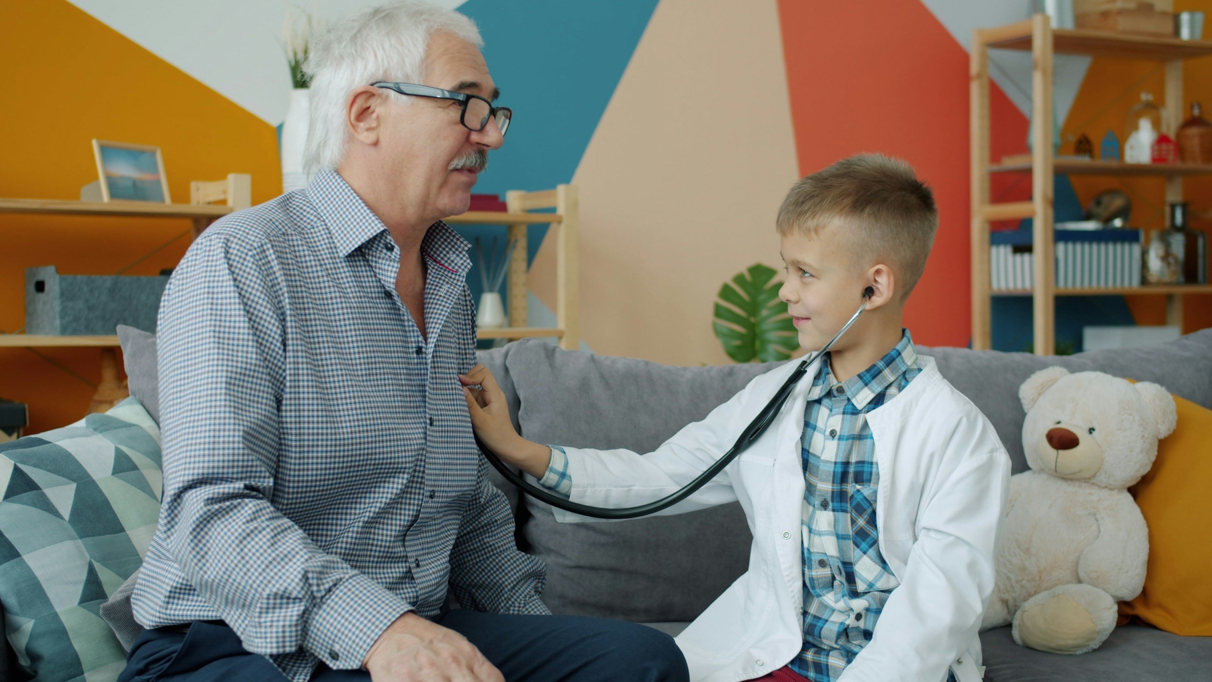 A young boy in a doctor's coat using a stethoscope on an elderly man sitting on a couch in a colorful living room.