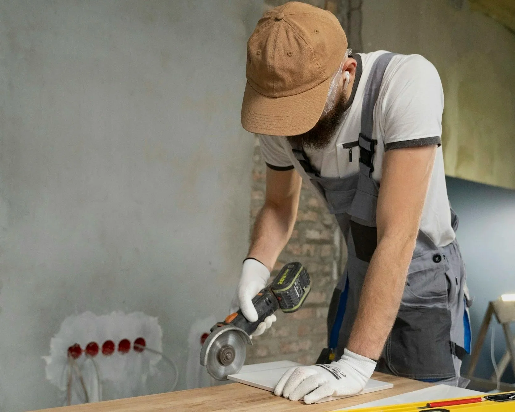 A man wearing a brown cap, white gloves, and grey overalls is using a power rotary tool to cut or sand a piece of white material on a wooden surface in a workshop setting.