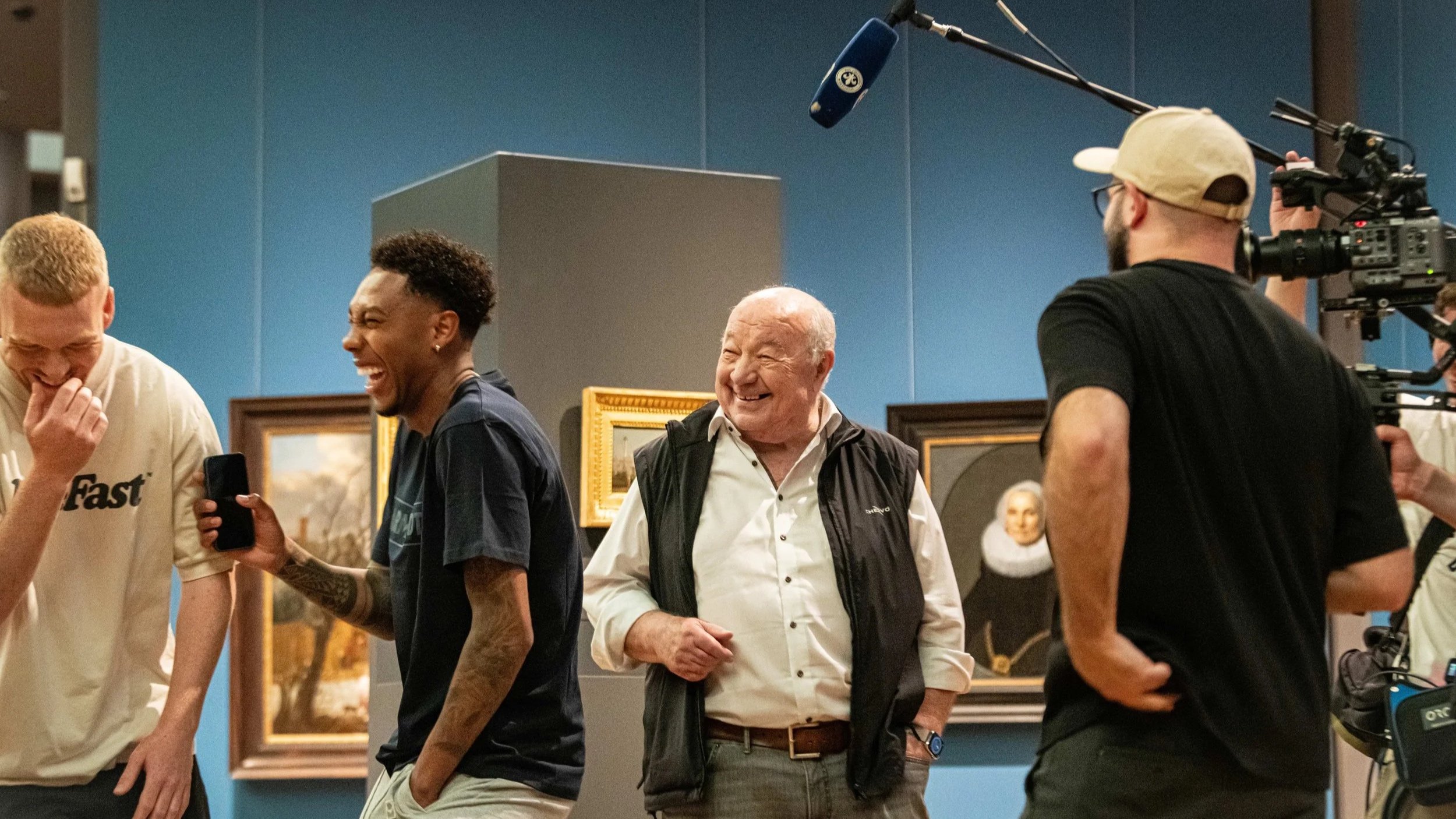 Menschen lachen und unterhalten sich in einer Kunstausstellung, im Hintergrund sind gerahmte Gemälde an blauer Wand.