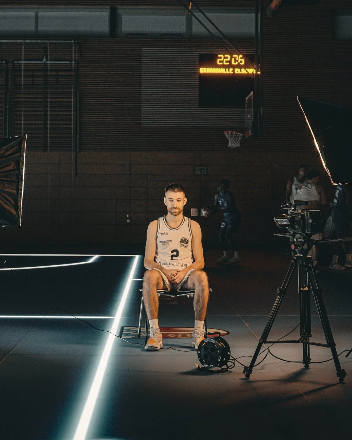 Ein junger Basketballspieler sitzt während eines Interviews auf einem Stuhl in einer Basketballhalle. Im Hintergrund sind weitere Spieler beim Spielen zu sehen, und es gibt eine Sportkamera sowie Beleuchtungsgeräte für Filmaufnahmen. Ein digitales Anzeigegerät an der Wand zeigt die Uhr und die Temperatur.