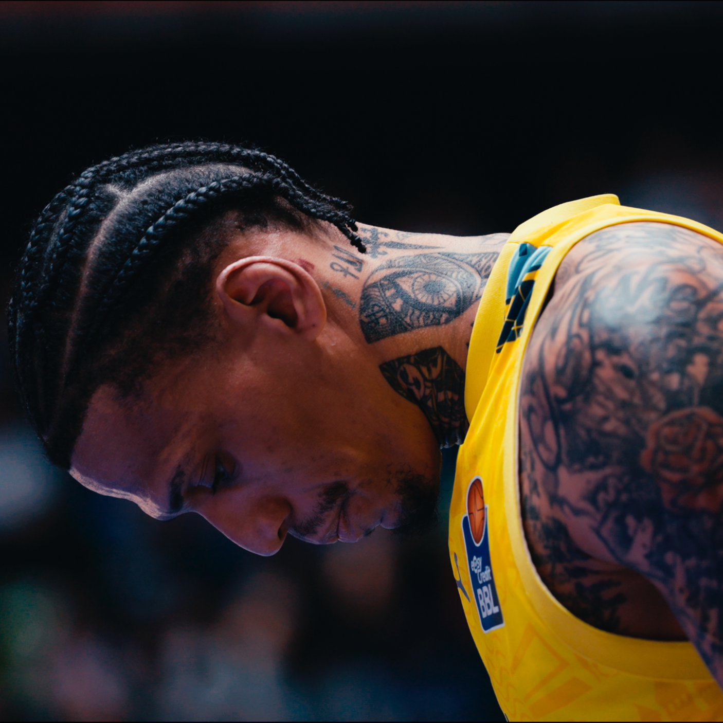 Nahaufnahme eines männlichen Basketballspielers mit Tattoos, der in einem gelben Trikot während eines Spiels nach unten schaut.