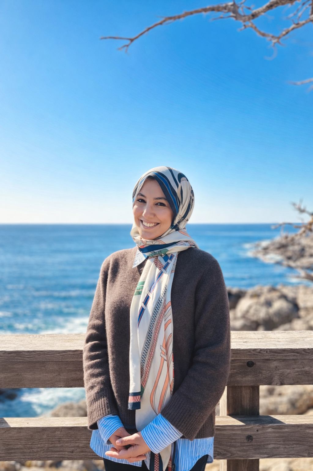 A woman with a patterned headscarf smiling by the ocean on a sunny day, standing near a wooden railing with rocks and the sea in the background.