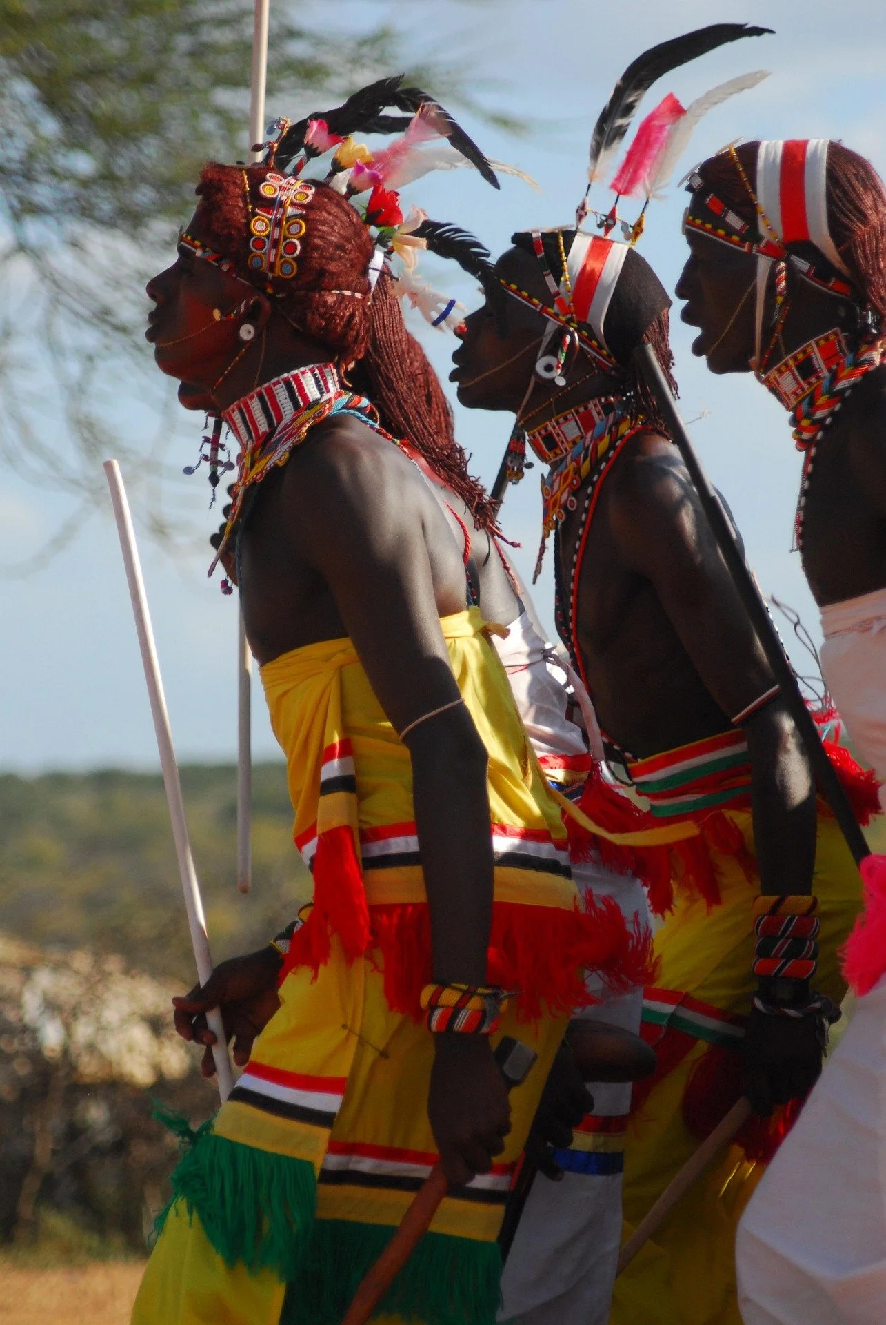 Three tribesmen dressed in colorful traditional attire with bead necklaces, feathers, and painted accessories, standing outdoors against a clear sky.