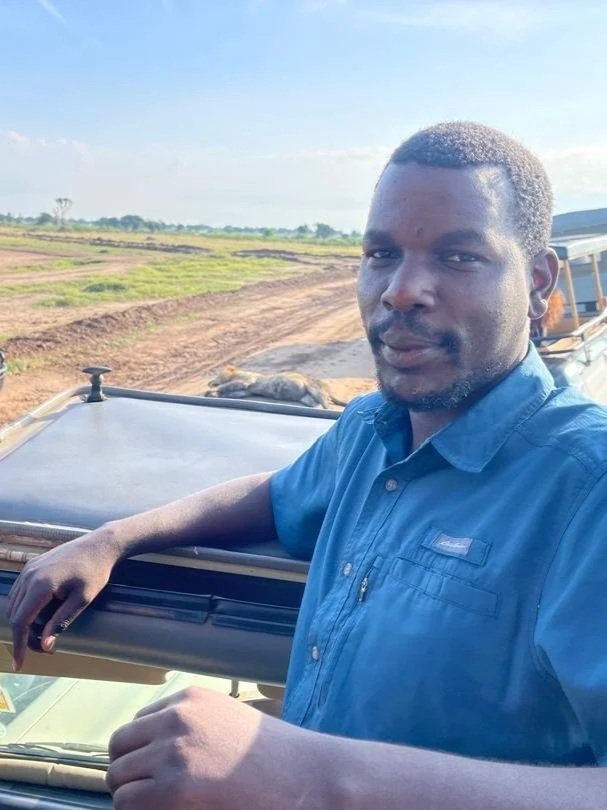A man in a blue shirt standing next to a vehicle on a dirt road in a rural area with fields and a cloudy sky in the background.