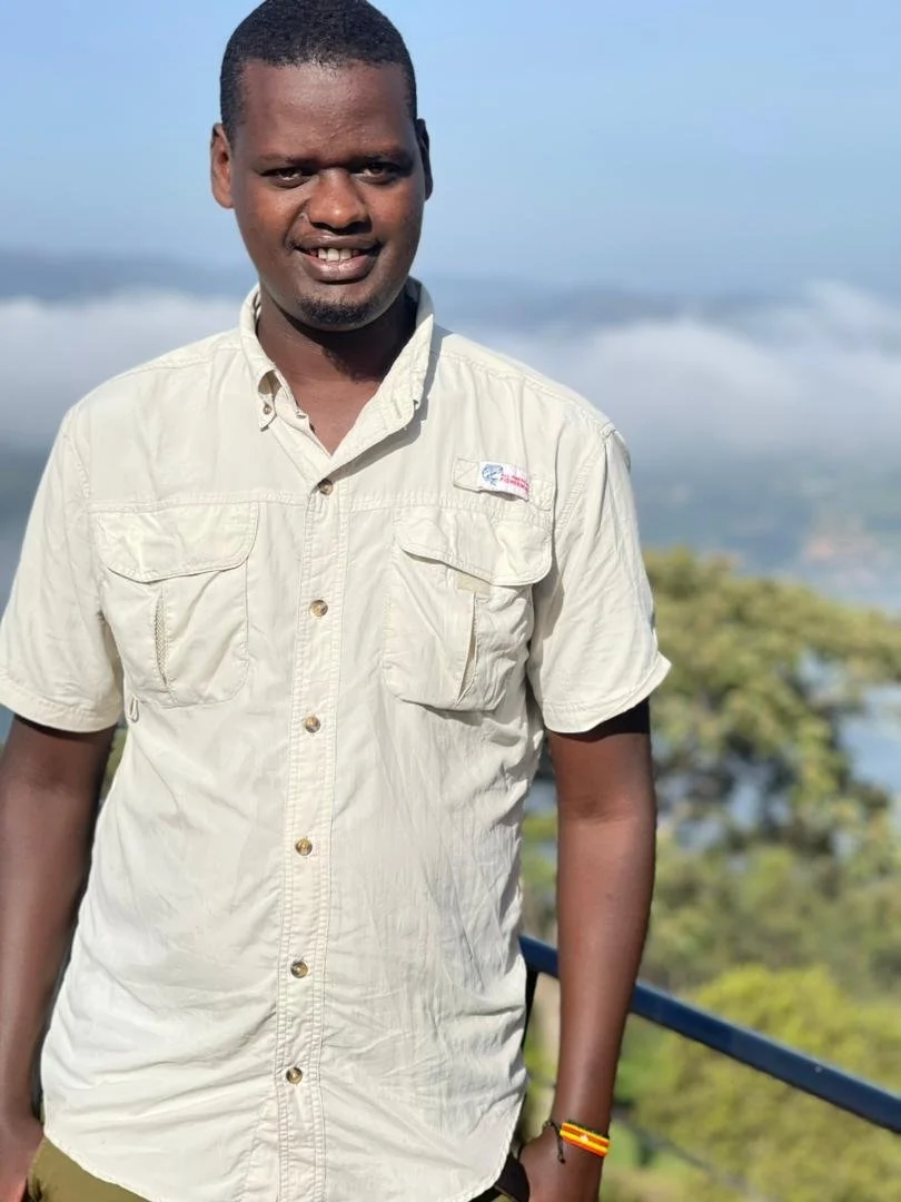 A man standing outdoors on a platform, smiling, with scenic mountains and trees in the background.