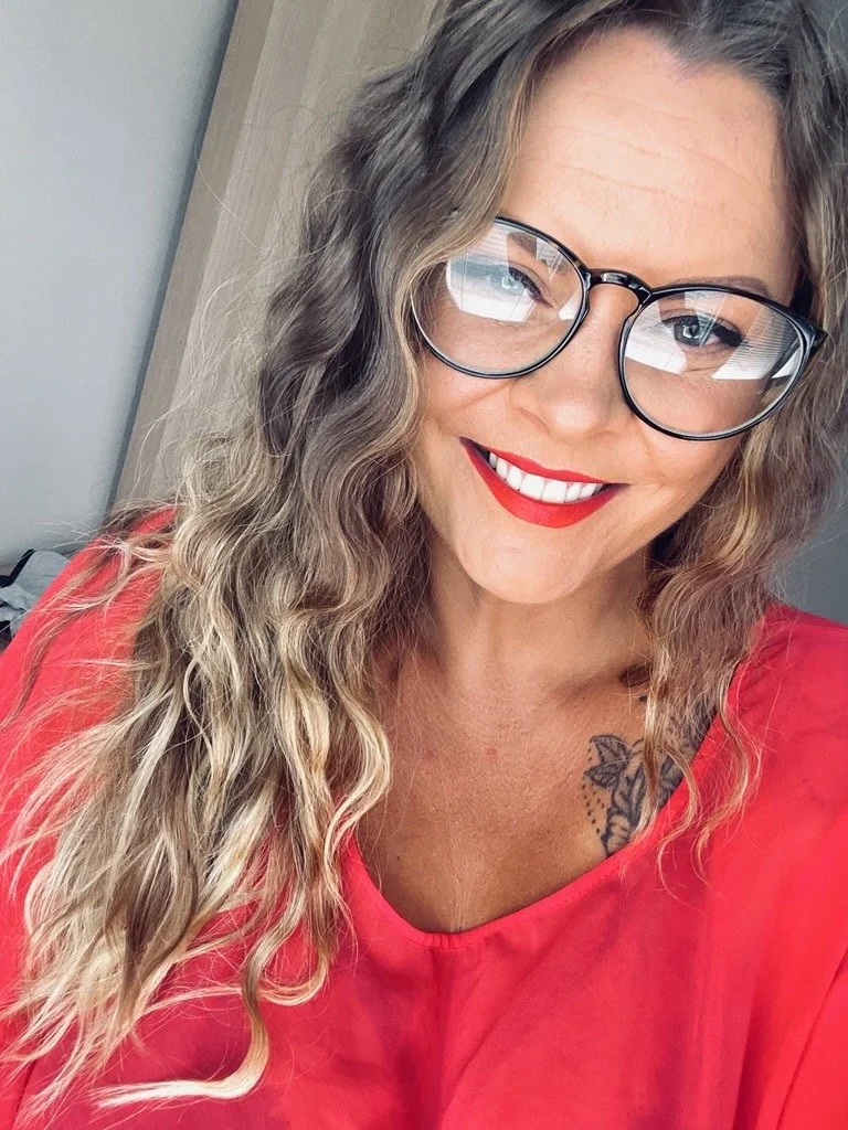 A smiling woman with wavy blonde hair, wearing glasses and a red top, showing a tattoo on her chest.