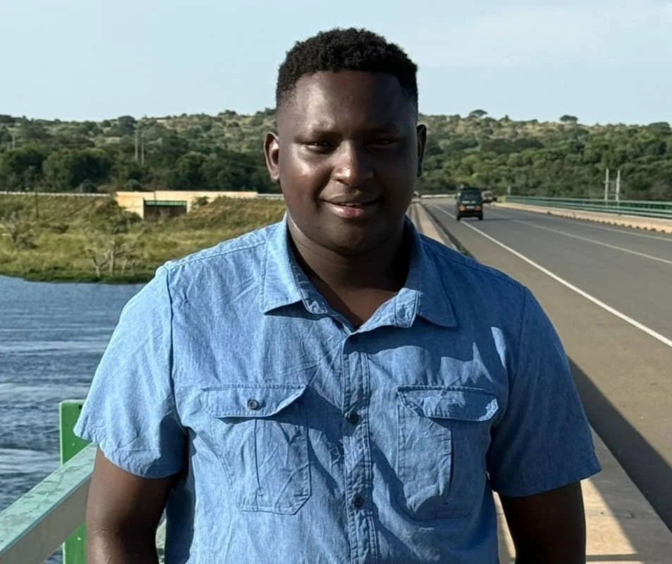 Young man standing outdoors near a body of water with a highway in the background.