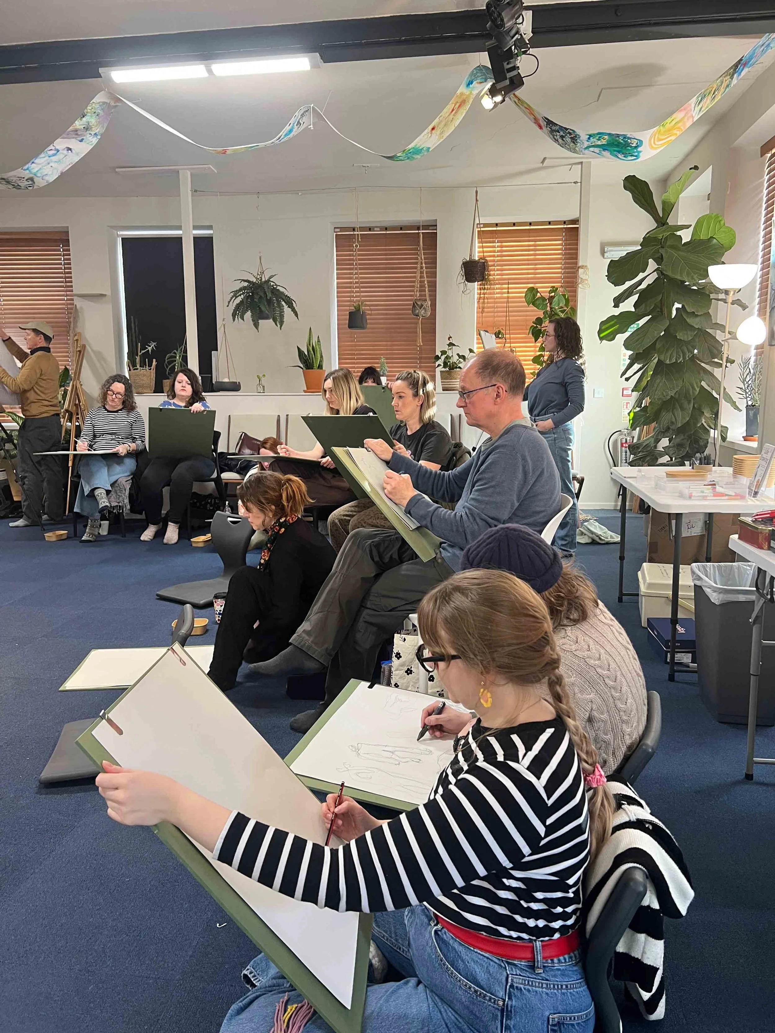 People sitting in a classroom drawing on large sketchpads, with indoor plants and decorative paper streamers hanging from the ceiling.