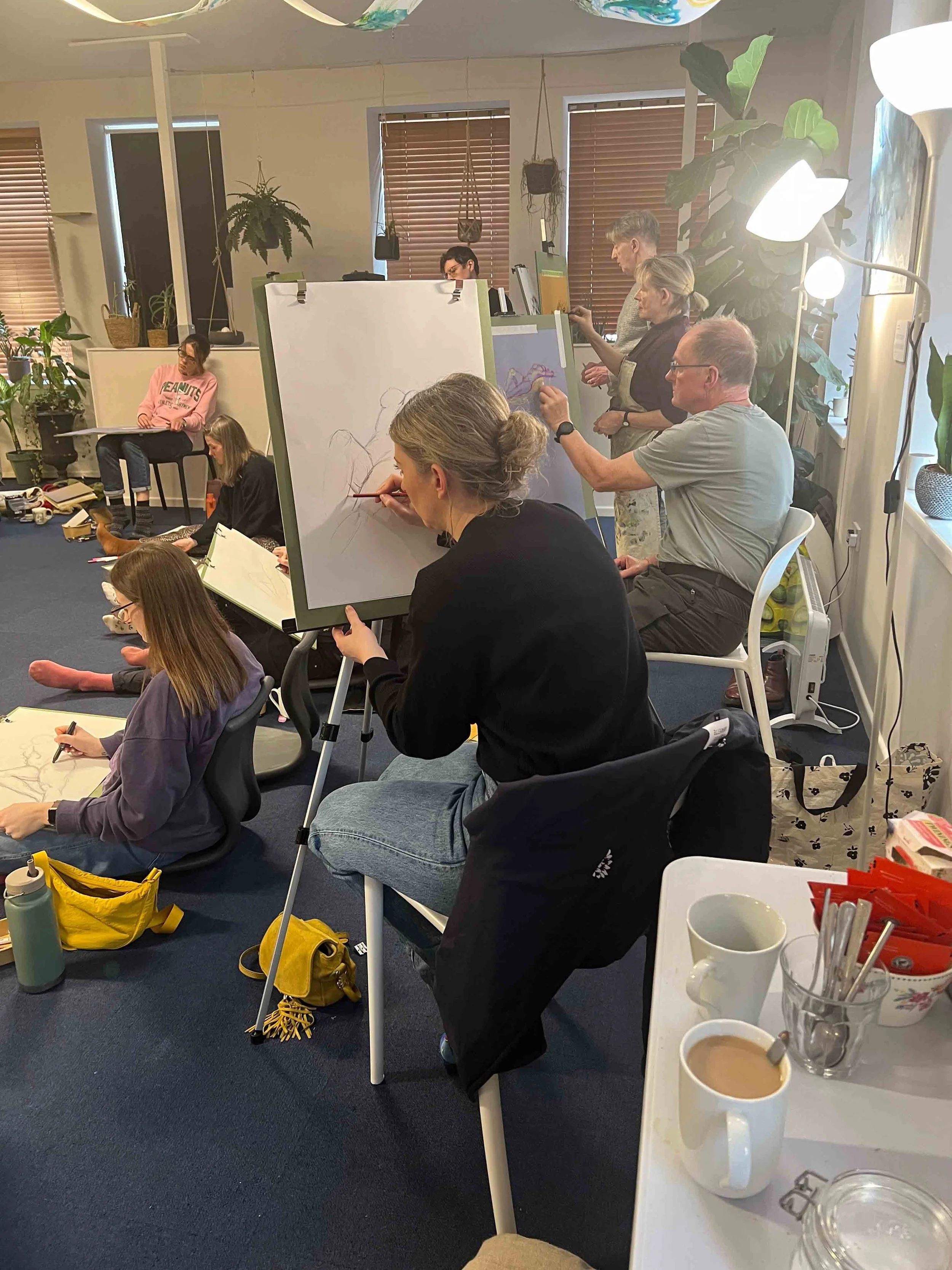 2People participating in an art class, drawing on paper and a whiteboard, with some sitting on the floor and others seated at desks, in a well-lit room with plants and windows.