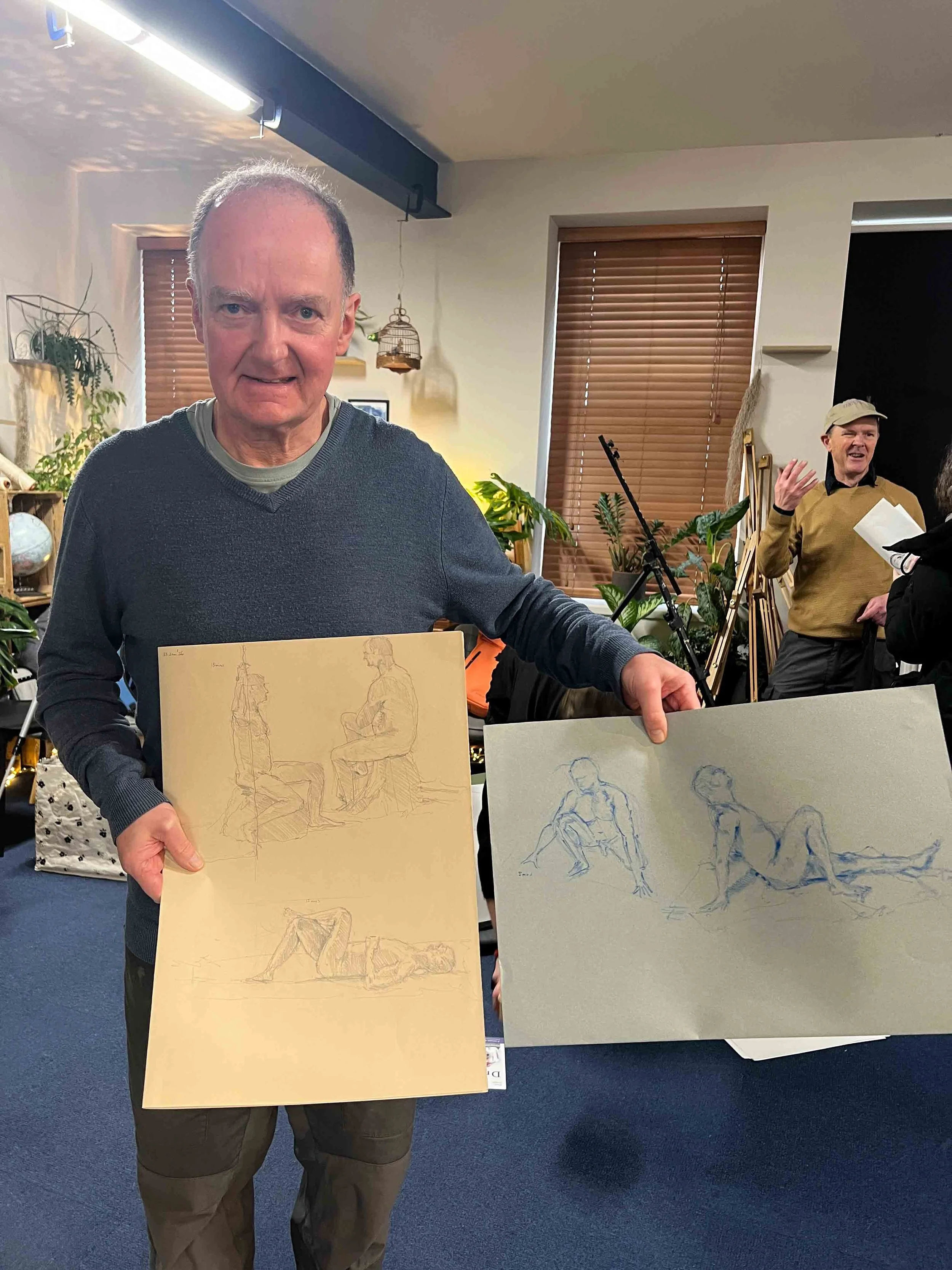 A man stands indoors holding two sketchbooks with figure drawings, smiling at the camera. In the background, other people are socializing, and there are plants, window blinds, and art supplies visible. Now, Draw!