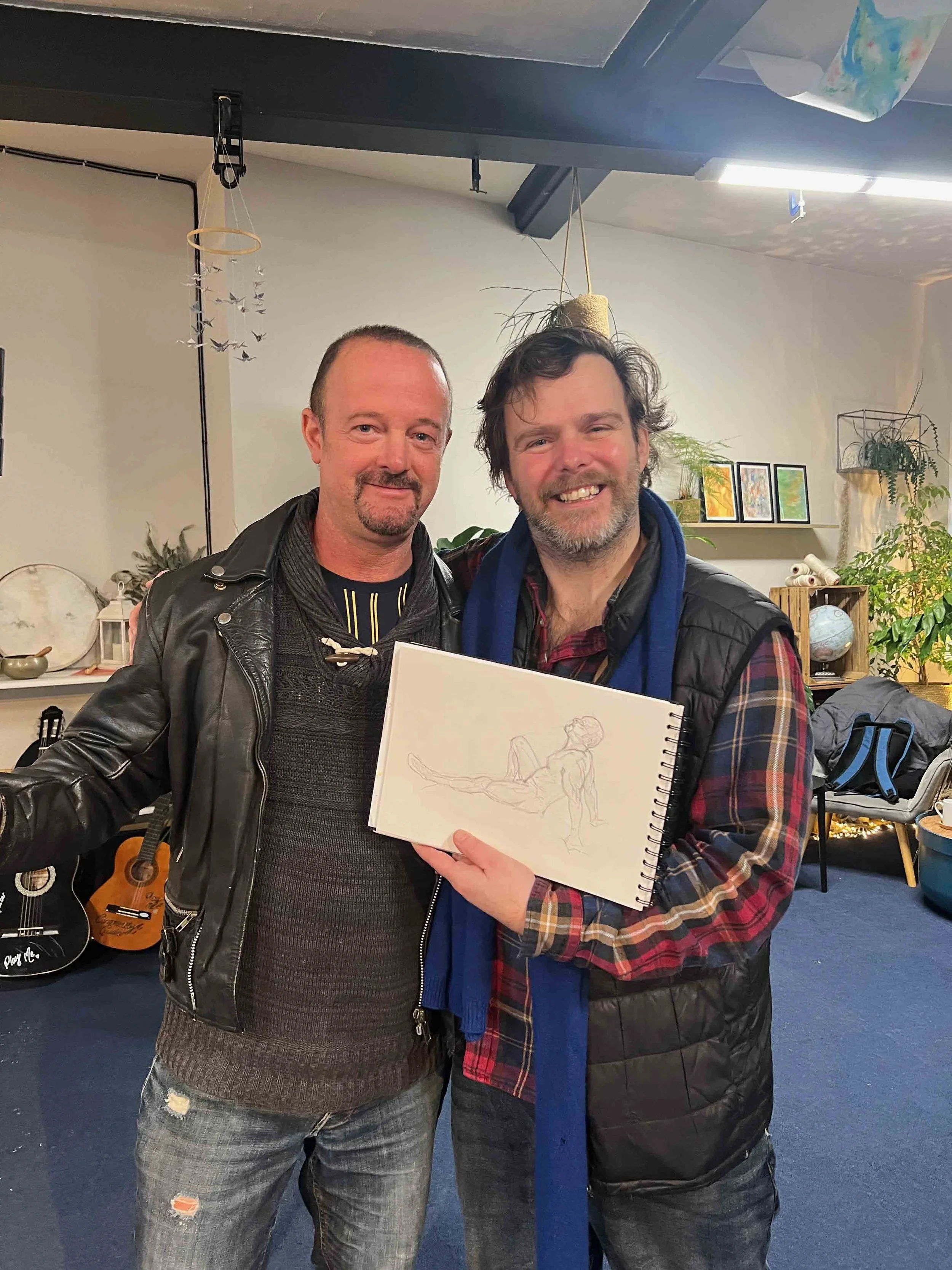Two men standing together, one holding a sketchbook with a figure drawing, in a cozy room decorated with plants, artwork, and musical instruments.