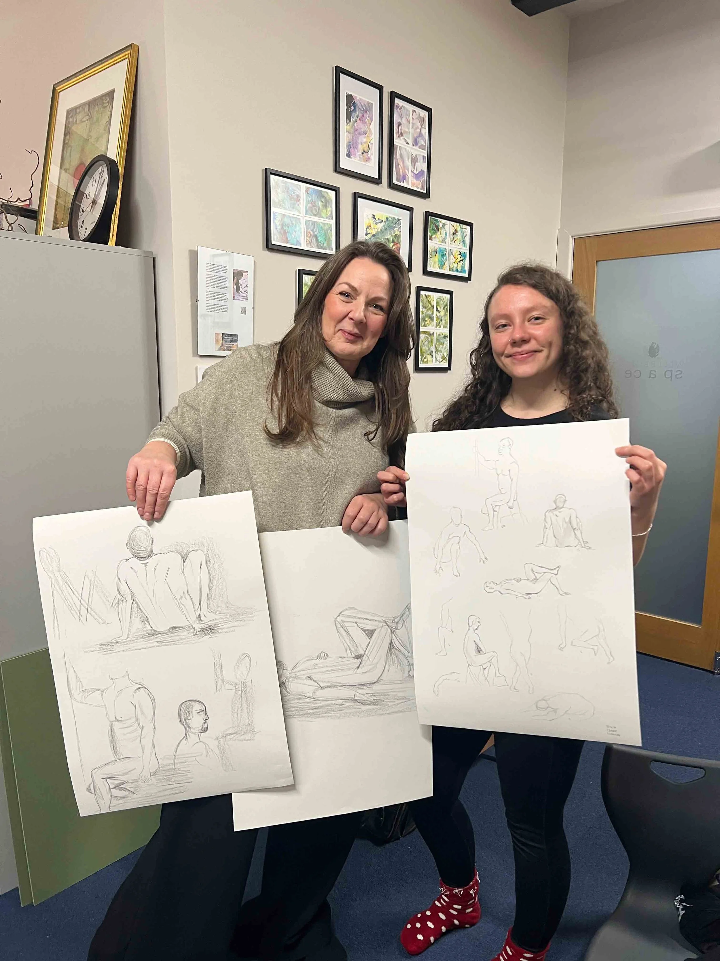 Two women standing in a room, holding sketch drawings of human figures, with framed artwork on the wall behind them. Now, Draw!