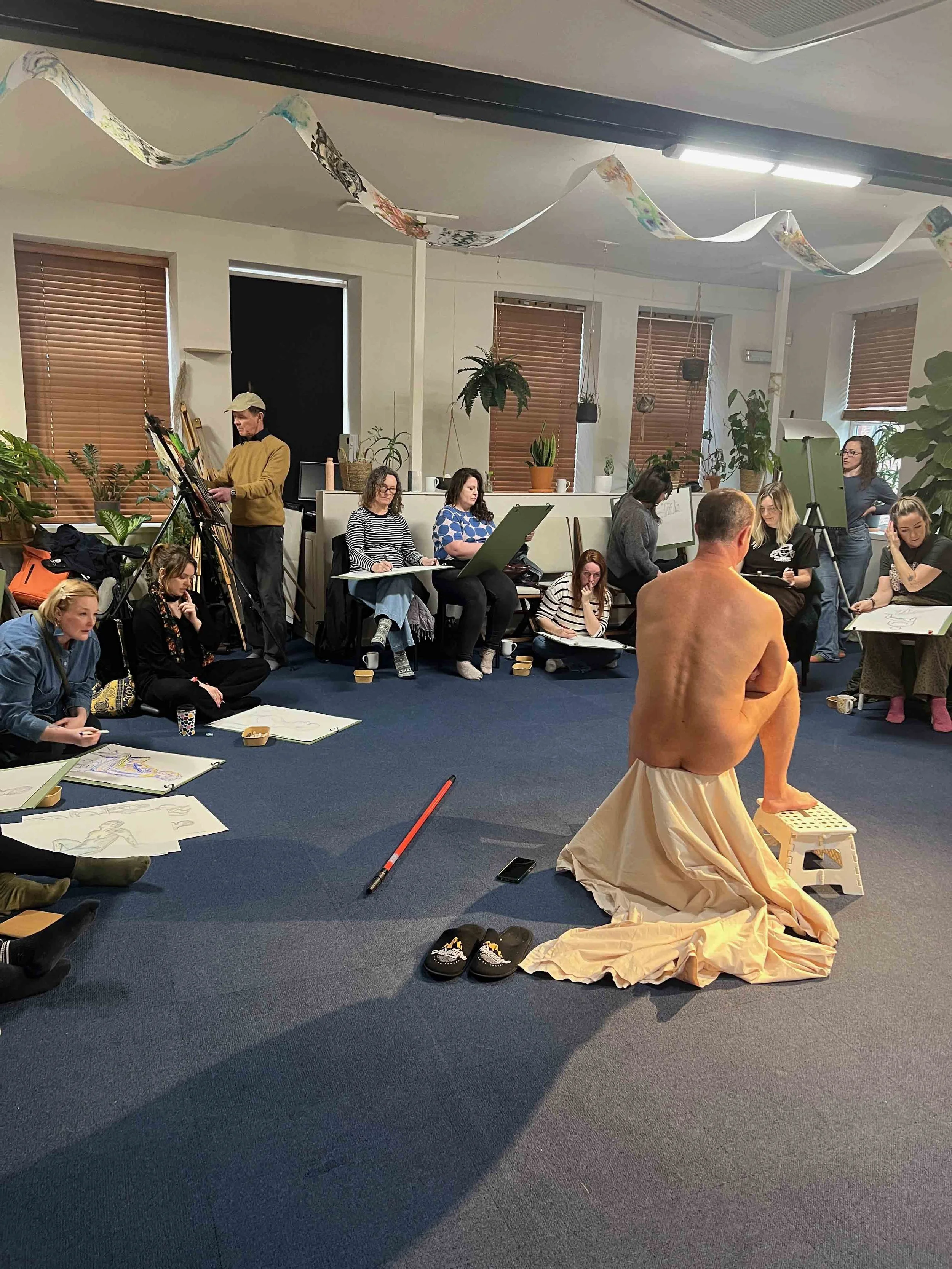 A group of people sitting on the floor and around the room, drawing or sketching on large sheets of paper. Life Drawing. Now, Draw!