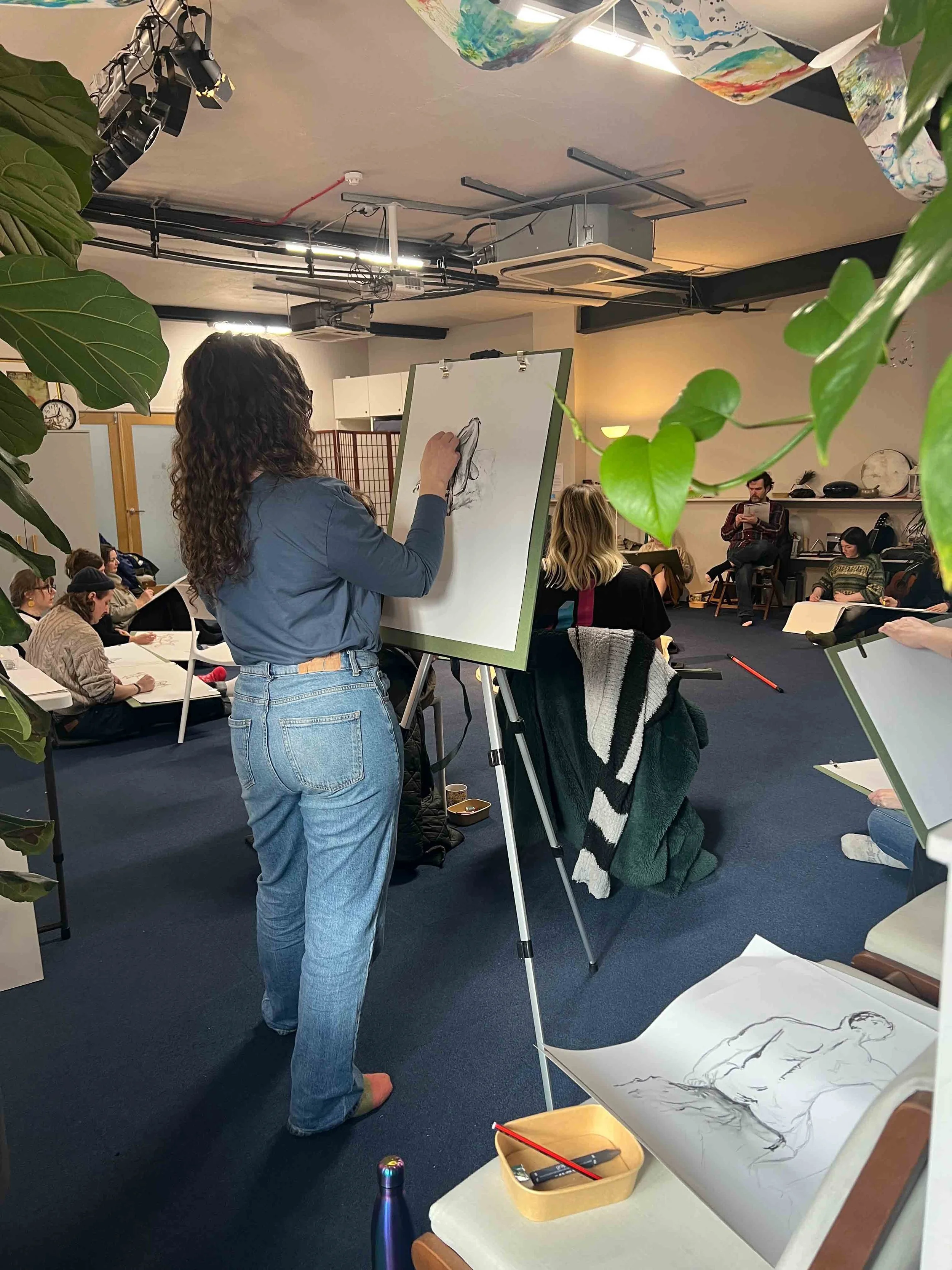 A woman with curly hair in a blue shirt and jeans drawing on an easel in a room full of seated people. Now, Draw! Macclesfield.