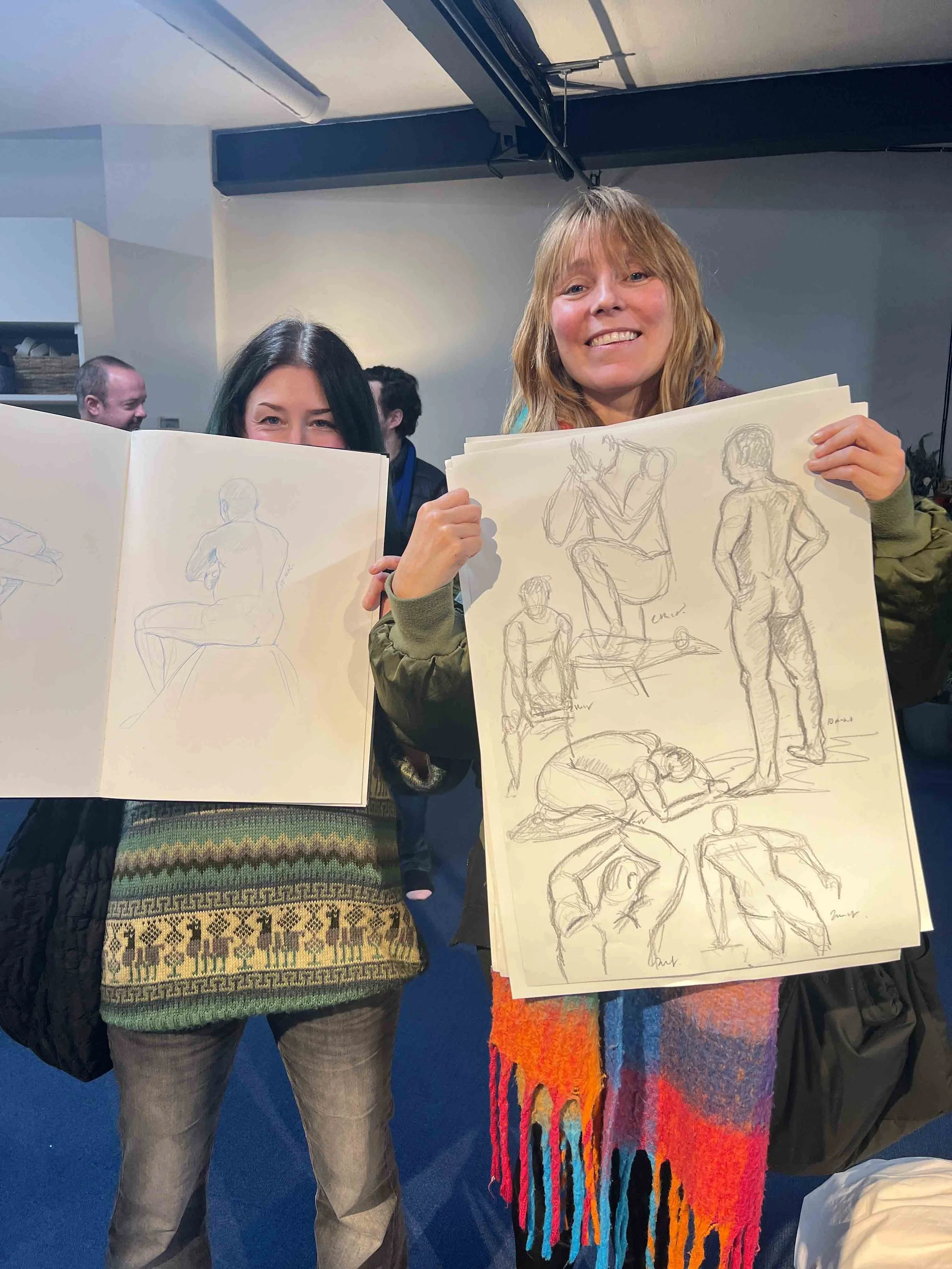 Two women smiling and holding large sheets of paper with figure sketches, standing indoors with other people in the background.