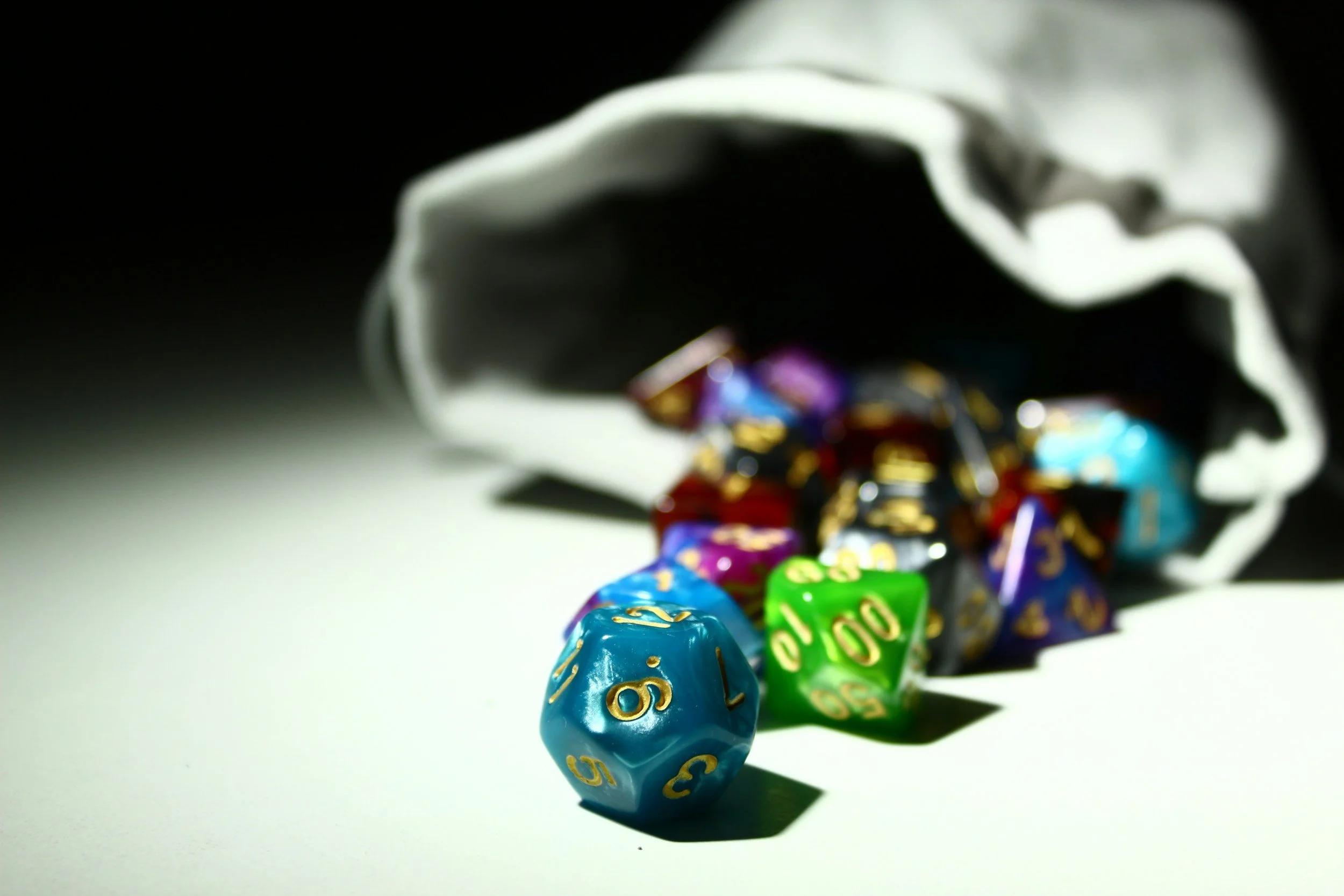Colorful polyhedral gaming dice spilled from a partially opened cloth bag on a white surface.