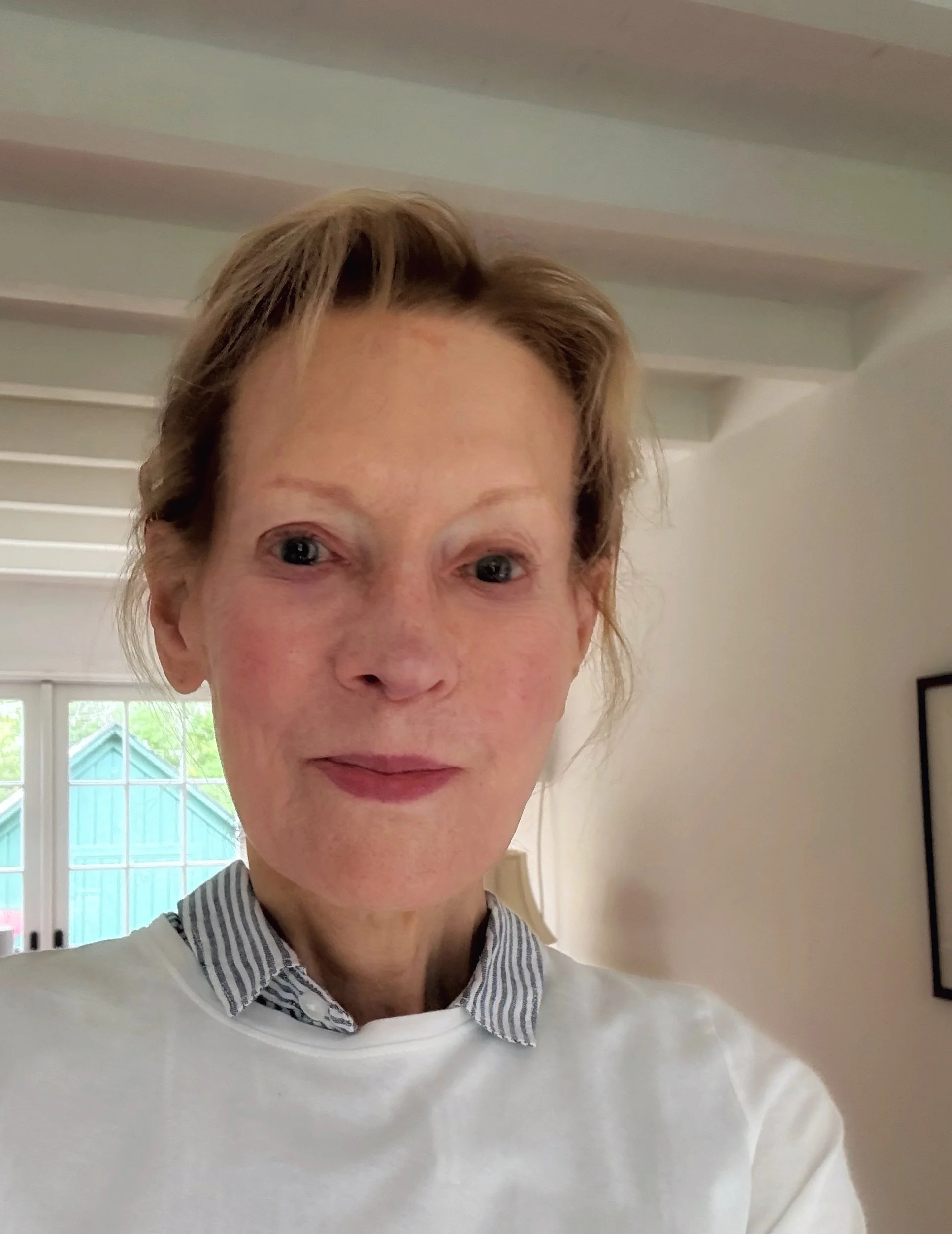 A close-up selfie of a woman with reddish hair, wearing a white top with a collared, striped shirt underneath, standing indoors with a window in the background.
