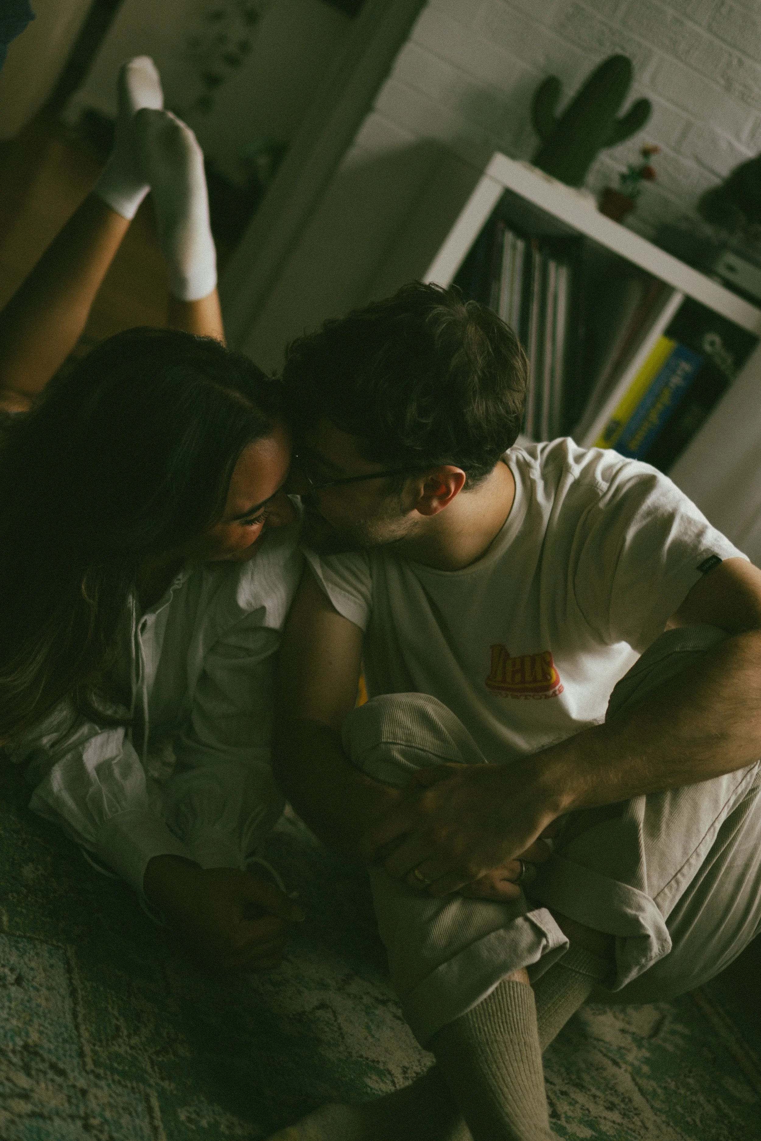 A man and woman are sitting on the floor, nose to nose, in a cozy indoor setting with a bookshelf and a cactus in the background.