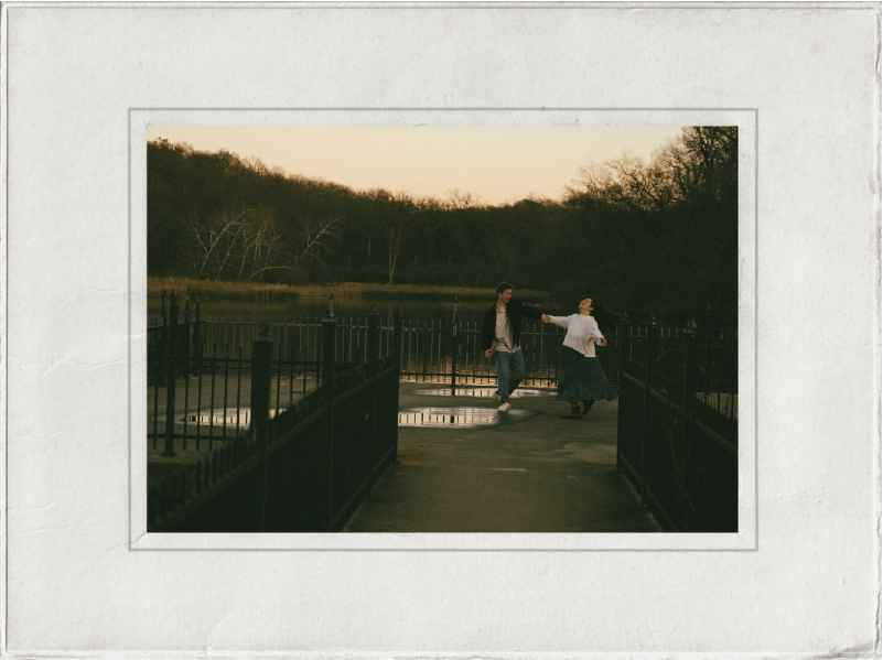 A couple holding hands and dancing on a bridge over a pond at sunset, surrounded by trees.
