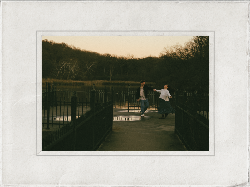 A couple holding hands and dancing on a bridge over a pond at sunset, surrounded by trees.