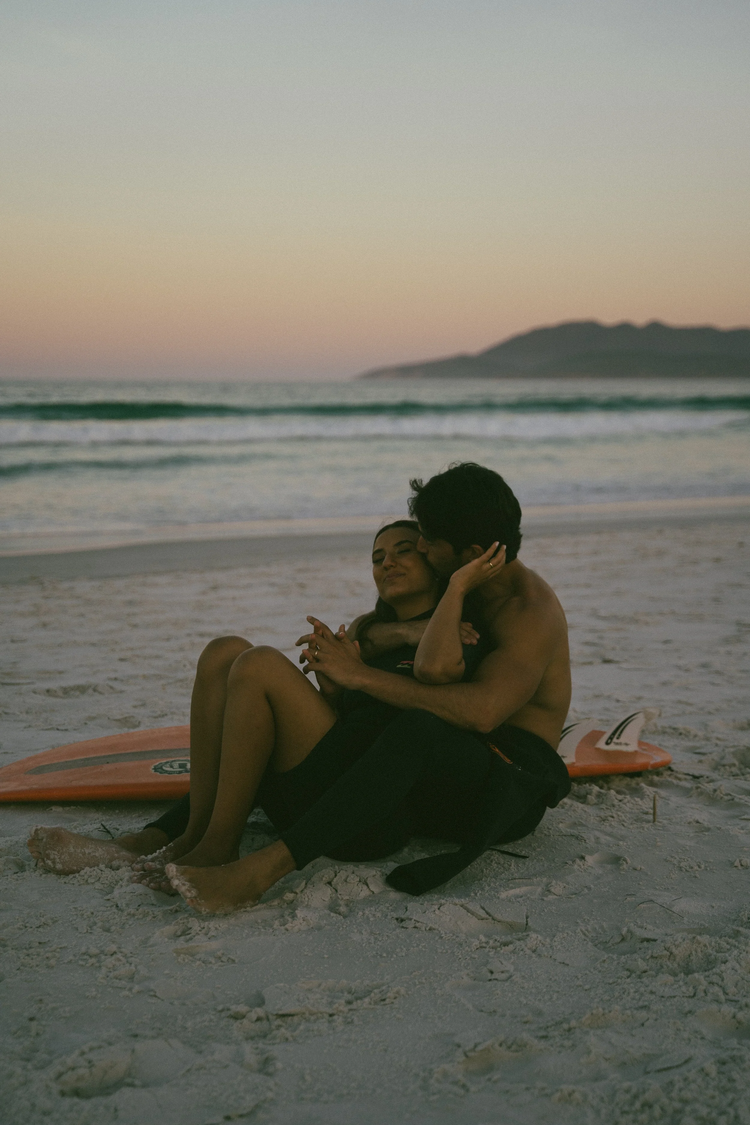 A couple sitting on the beach at sunset, embracing each other with ocean waves and a distant hill in the background.