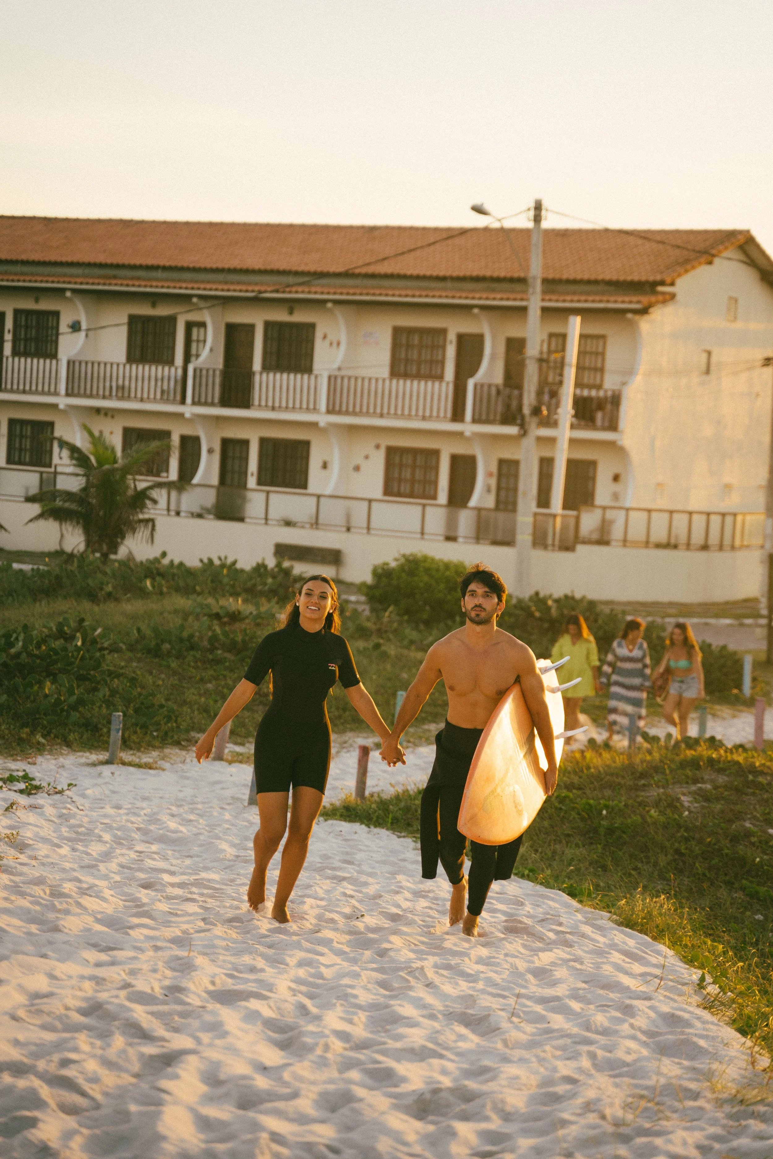 A man and woman holding hands and walking barefoot on the beach during sunset, with surfboards under their arms, and a group of people walking behind them.