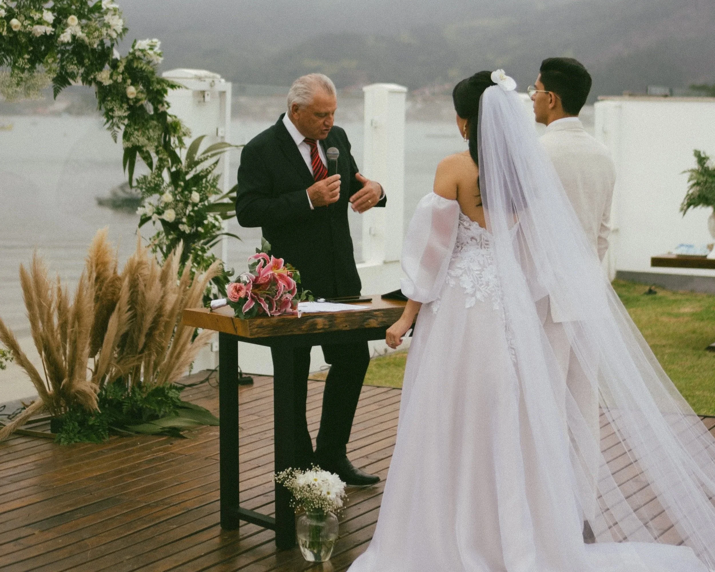 A wedding ceremony taking place outdoors by the water with a bride and groom standing in front of a man reading vows, surrounded by floral decorations.