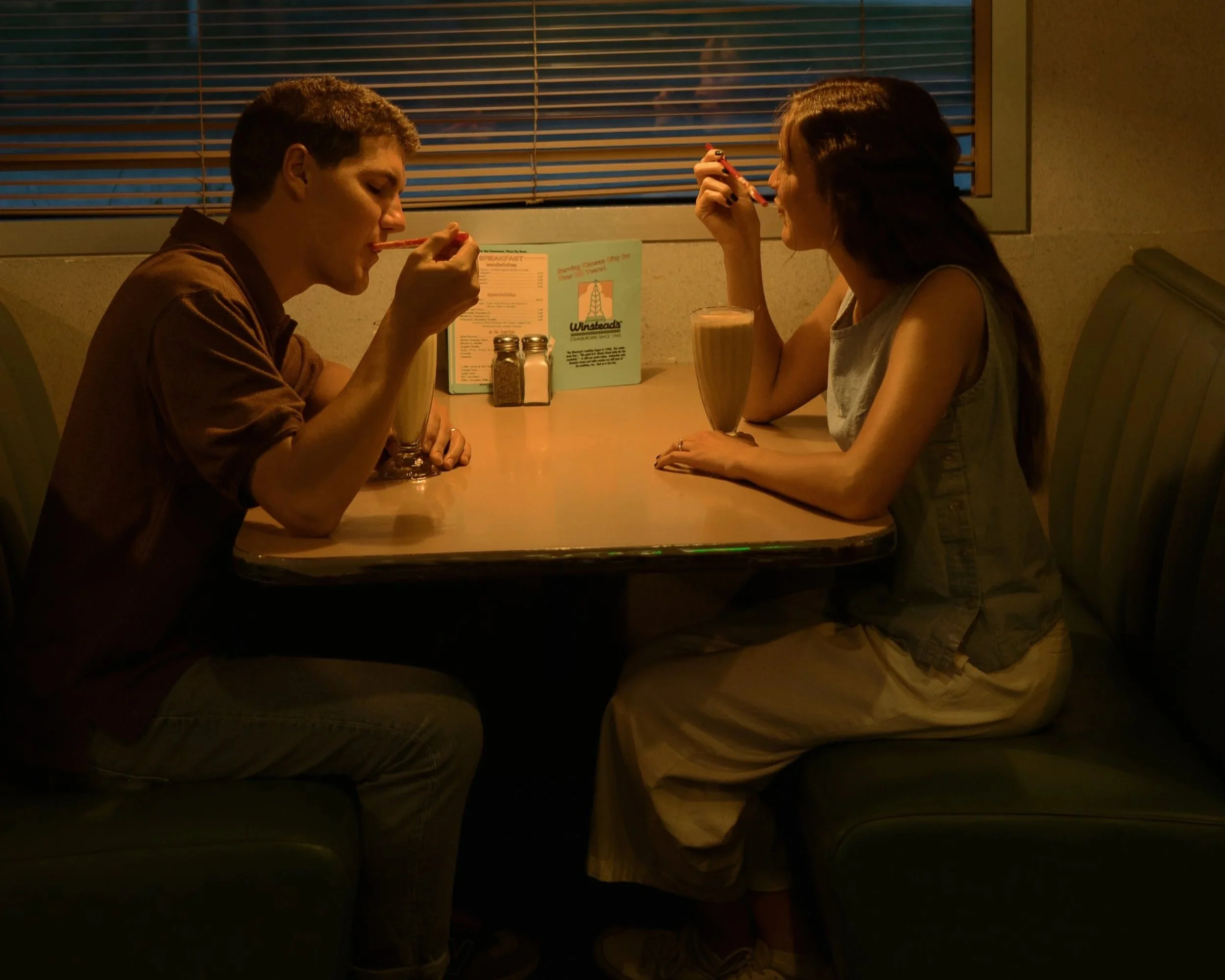 A young man and woman sit across from each other in a diner, each enjoying a milkshake and using a spoon.