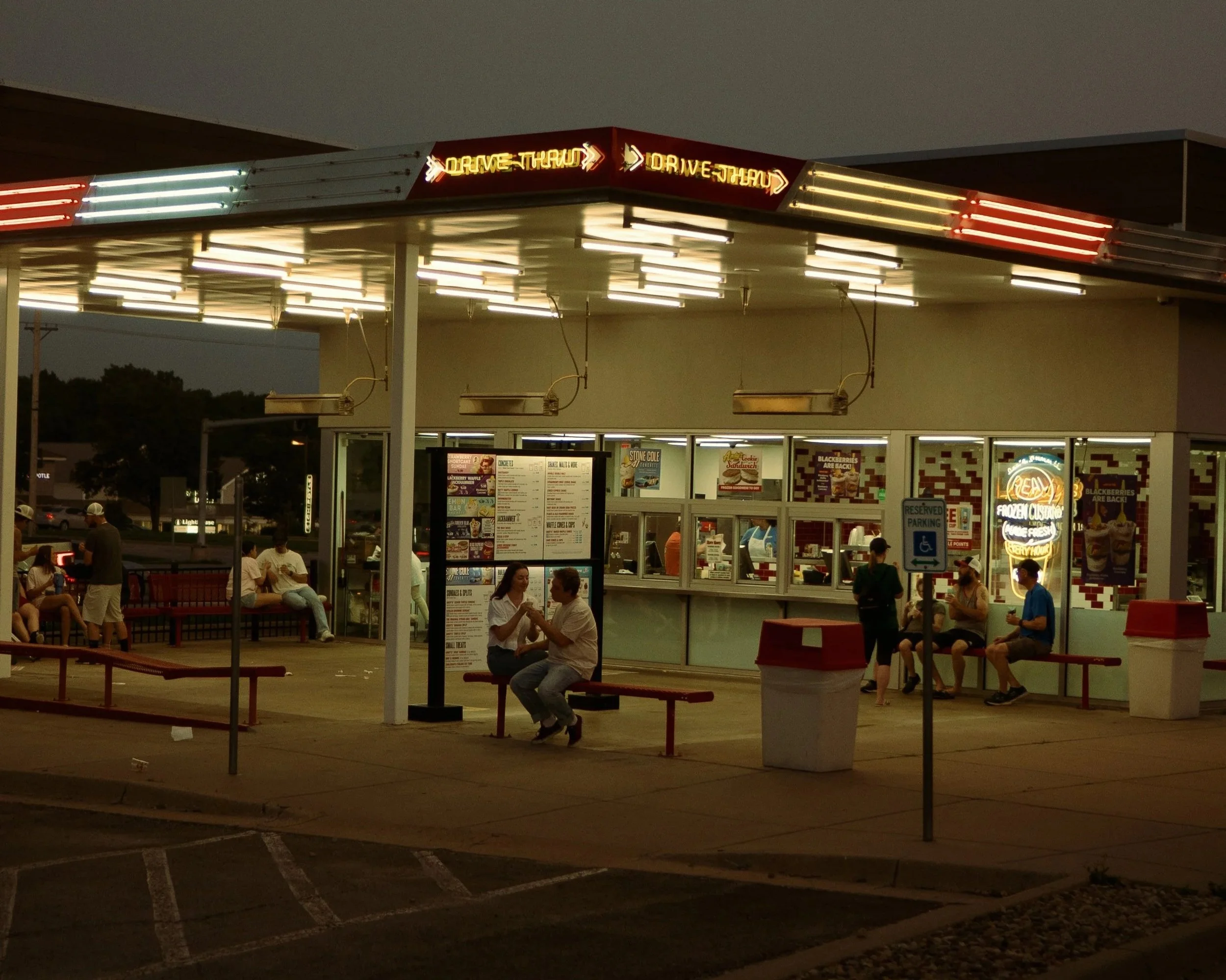 A night scene of a fast-food restaurant with a drive-thru, illuminated neon signs, and several customers sitting on red benches outside the building.