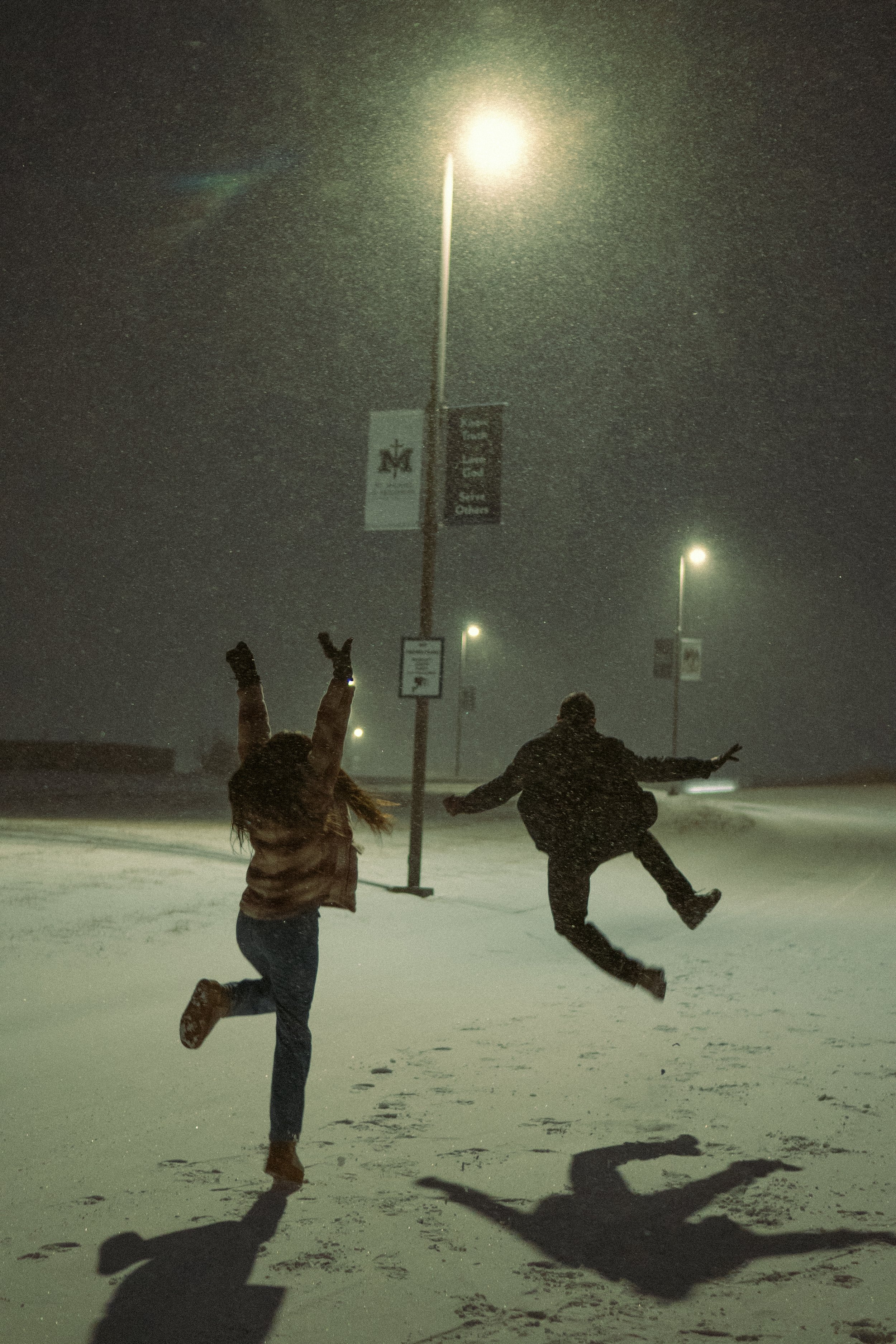 Two people playfully jumping in the snow on a dark, snowy night under streetlights, with a lamppost and banners visible in the background.