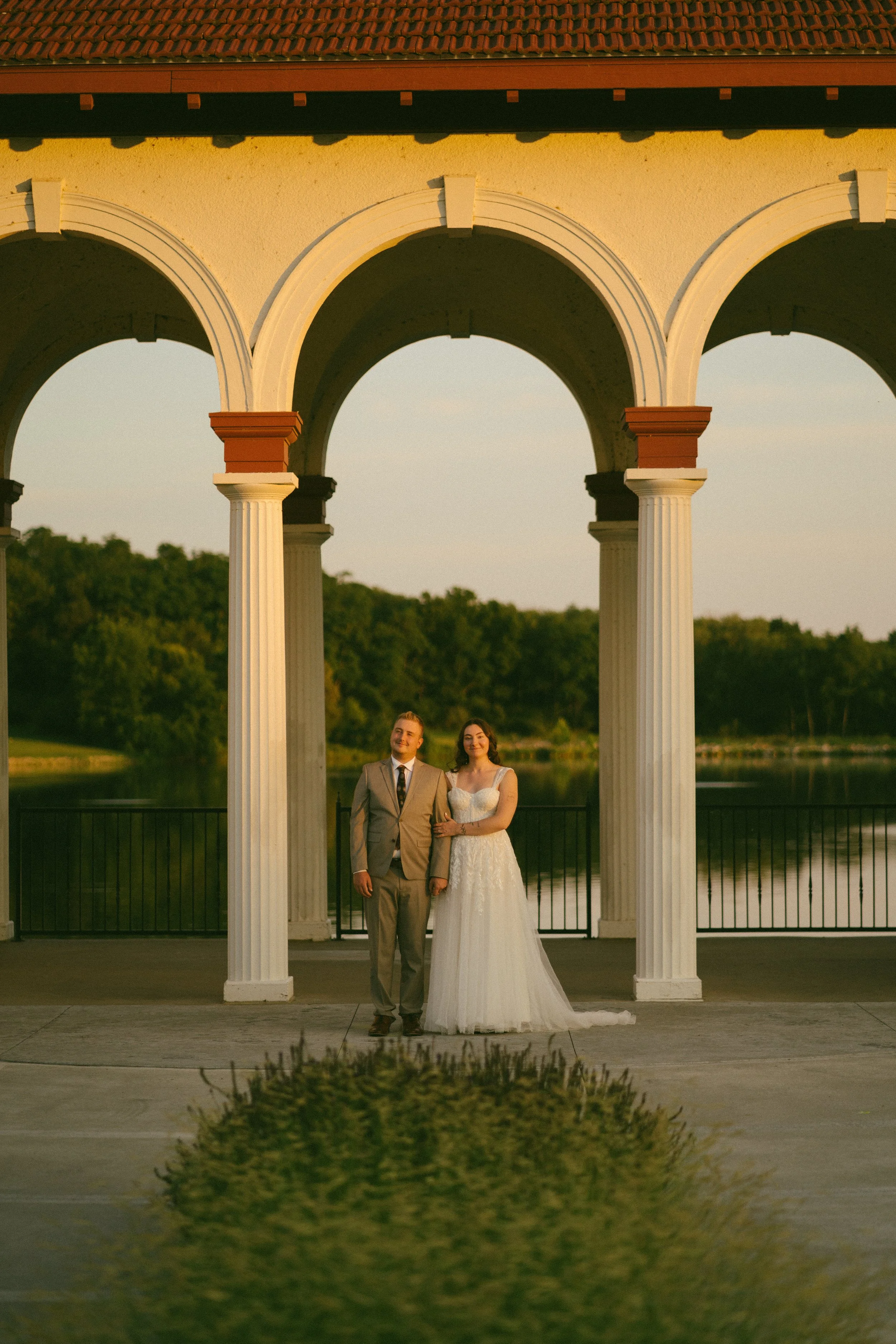A newlywed couple standing under a white arched structure with a lake and green trees in the background during sunset.