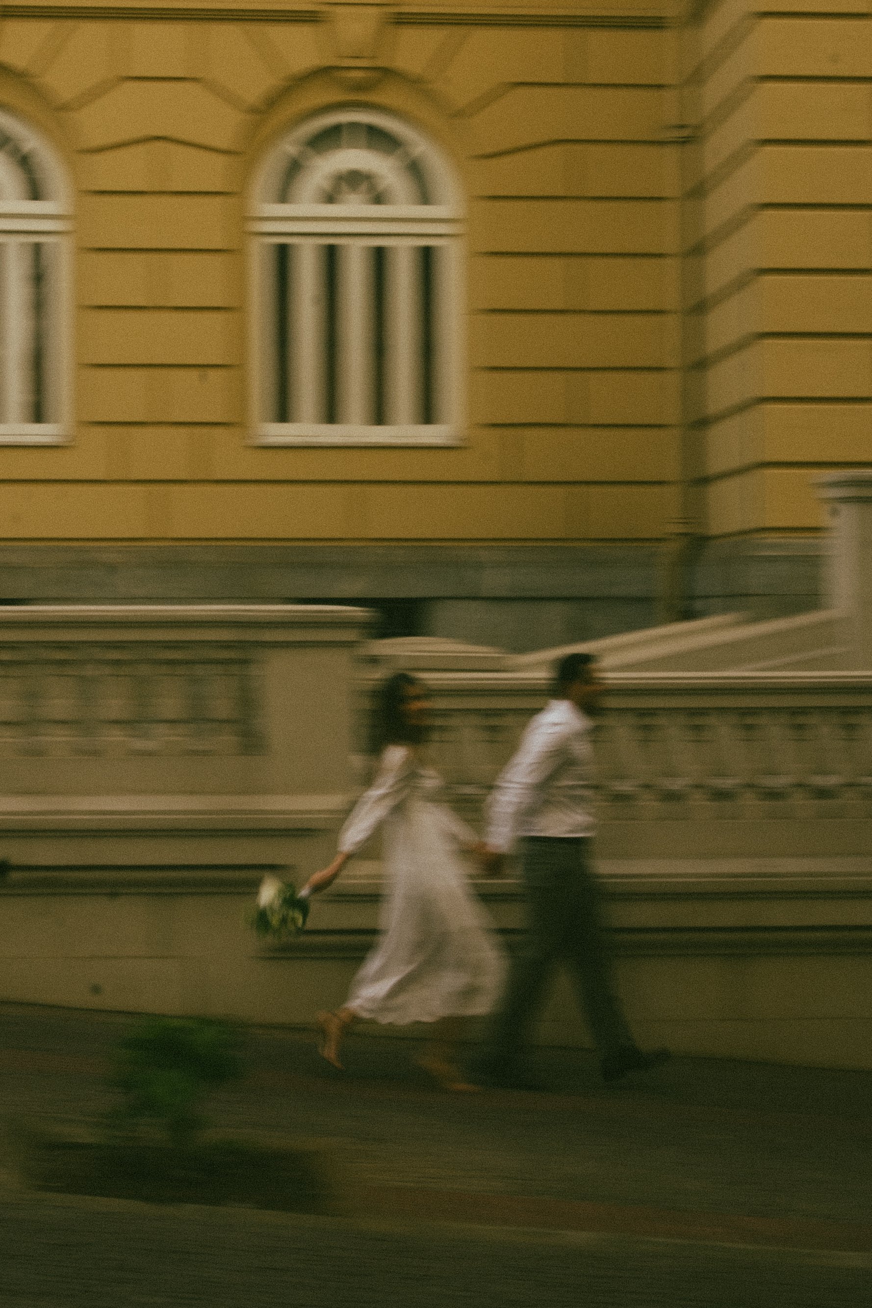 A blurred photo of a couple walking hand in hand on a city sidewalk at night, with a yellow building with arched windows in the background.