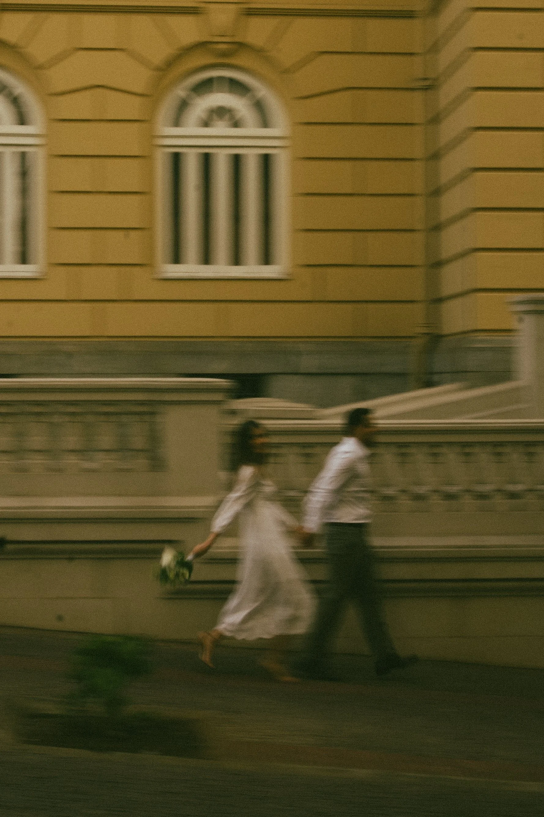 A blurred photo of a couple walking hand in hand on a city sidewalk at night, with a yellow building with arched windows in the background.