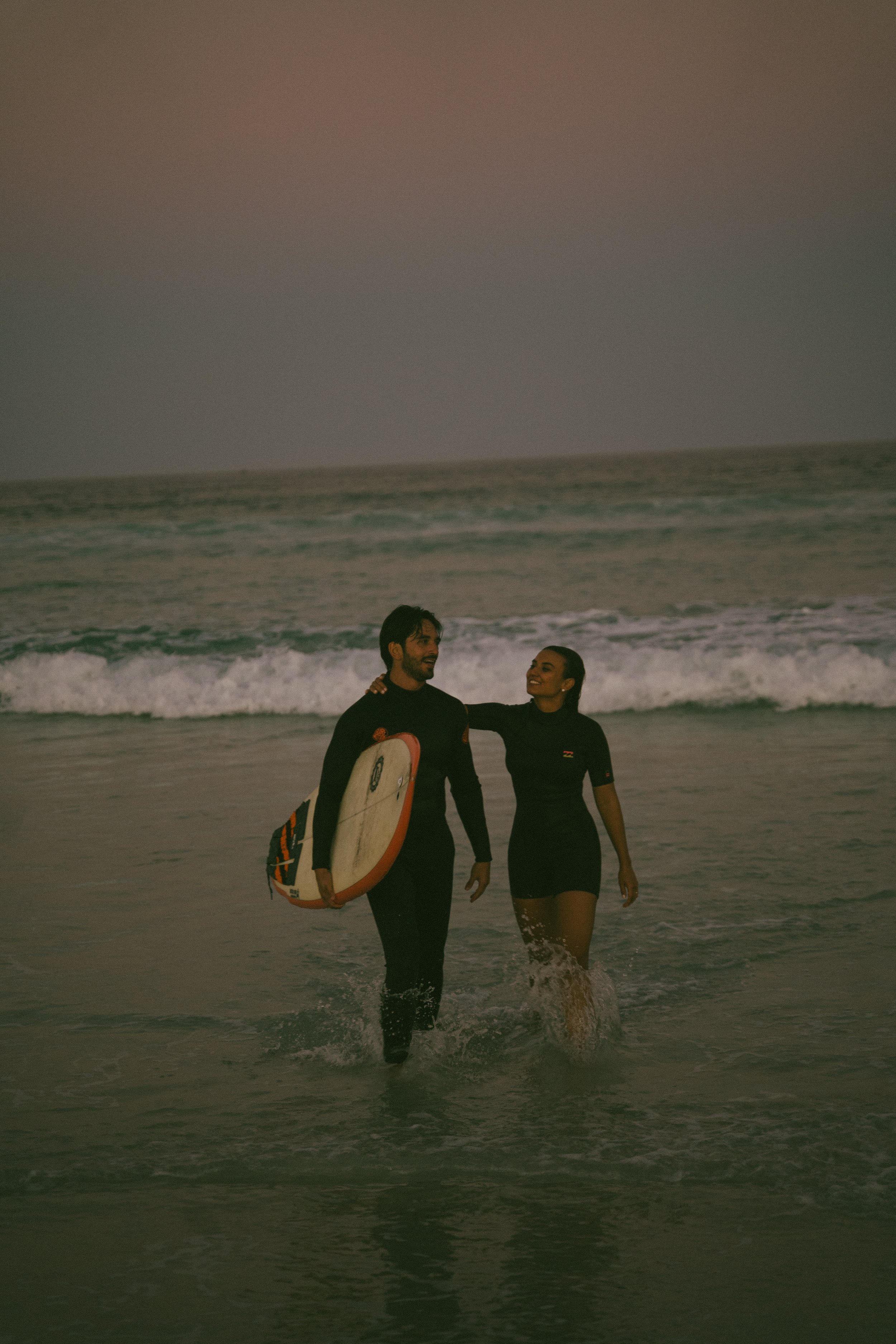 A man and woman walking in the ocean with a surfboard, smiling and looking at each other during sunset.