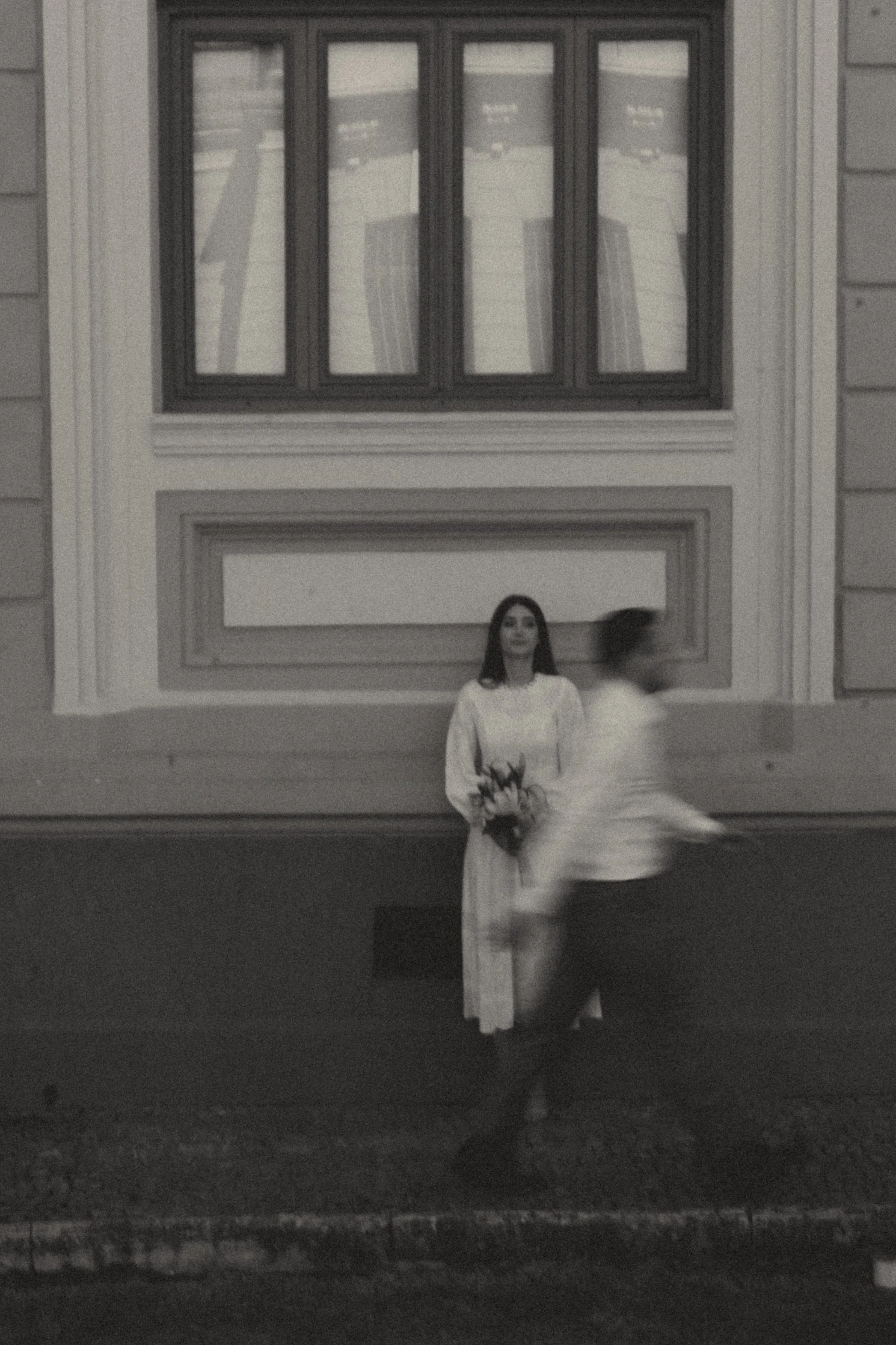 A woman in a white dress holding flowers standing against a wall with a window above her, as a person in motion passes by in front of her.